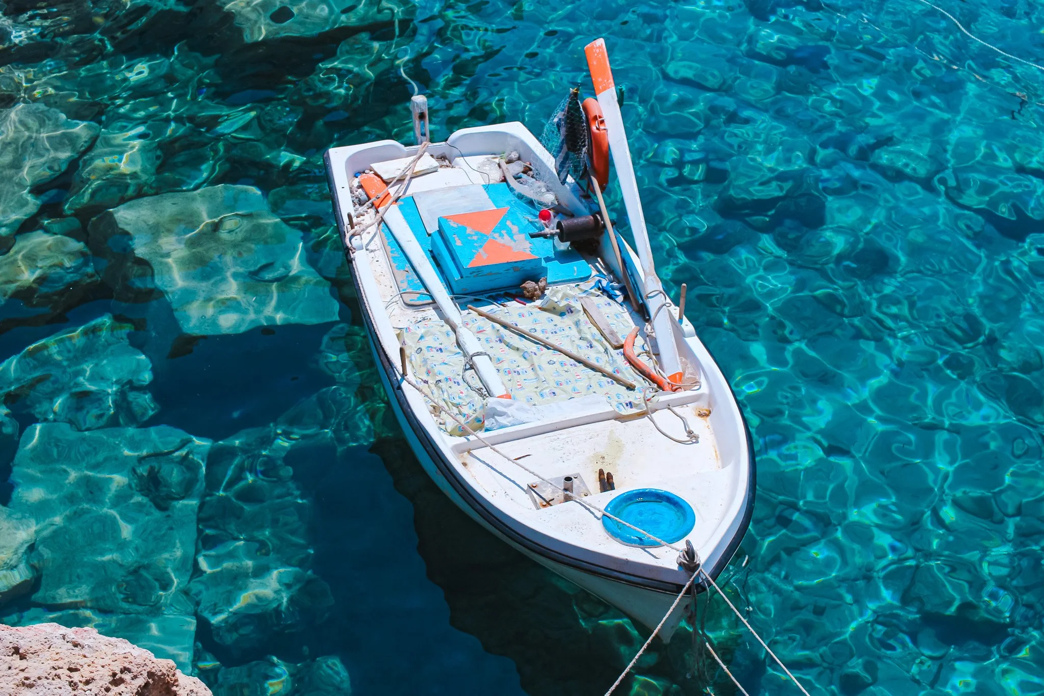 A small white boat with various objects inside floats on clear blue water near rocks.