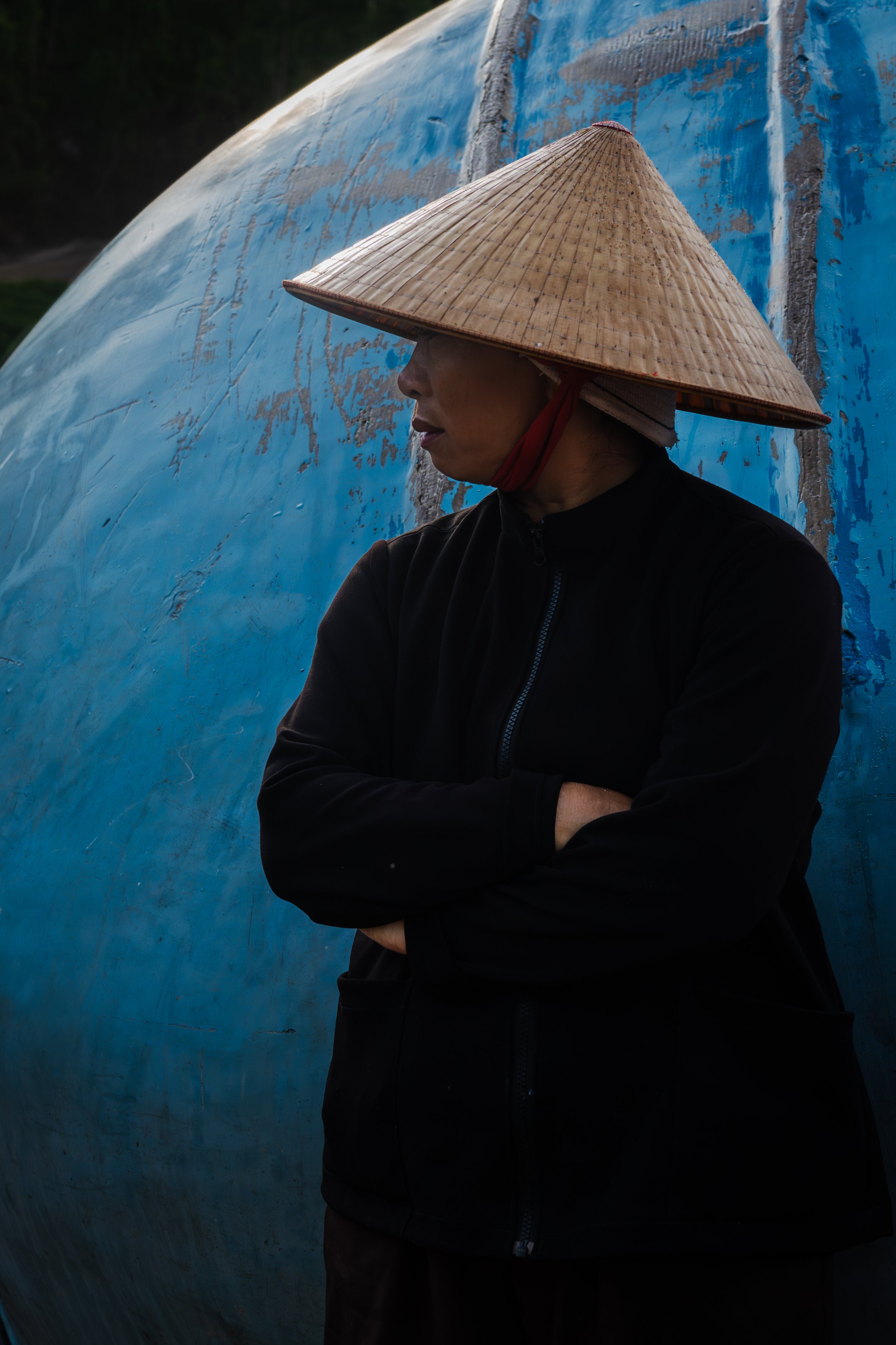 A Vietnamese women wearing a traditional conical hat with arms crossed, standing against a weathered blue boat in a fishing village.