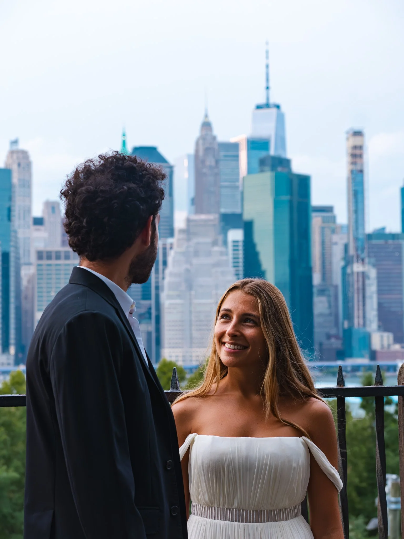 A groom in a black suit looking at a bride in a white dress, smiling at each other, with the New York City skyline in the background.