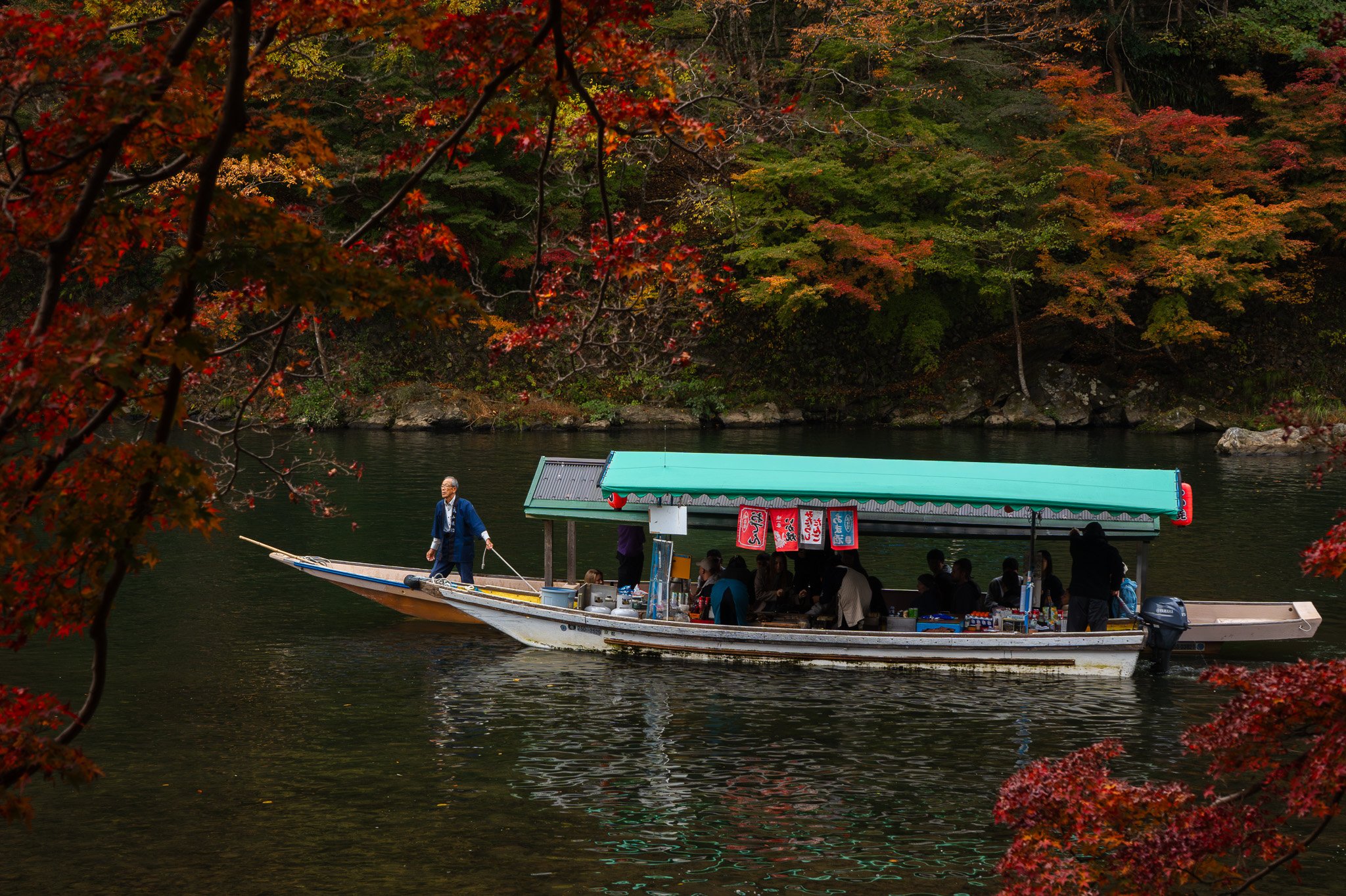 A boat with a green canopy floating on the Arashiyama river during autumn, with red and orange foliage in the background in Kyoto.