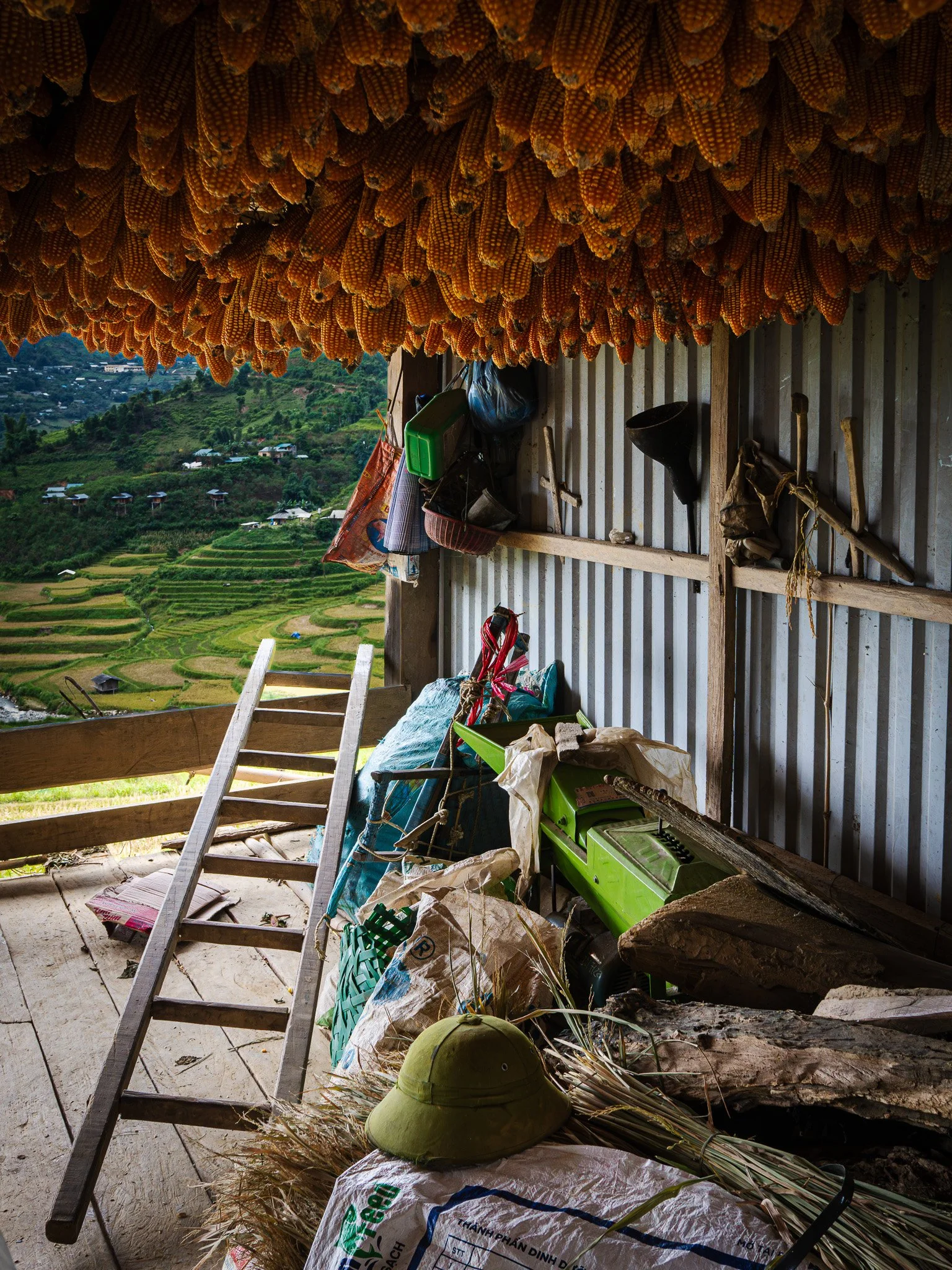 Inside view of a rustic wooden shelter with rows of dried corn hanging from the ceiling, a green vintage sewing machine, a wooden ladder, various bags, and household items, overlooking a vibrant terraced landscape with gardens and hills.