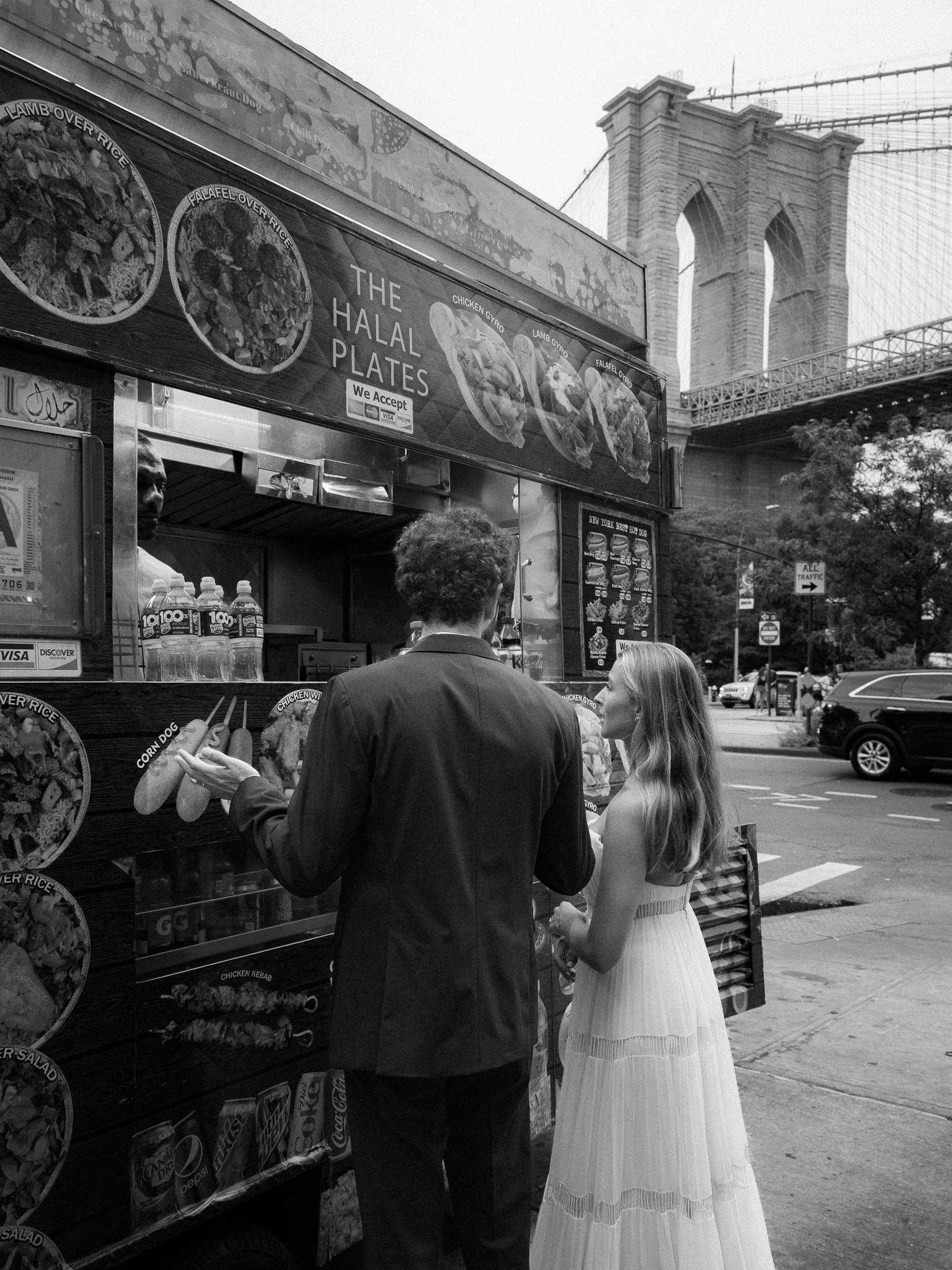 A bride and groom placing an order at a food cart in front of the Brooklyn Bridge in New York City, with a sign advertising halal plates and various food options, shot in black and white.