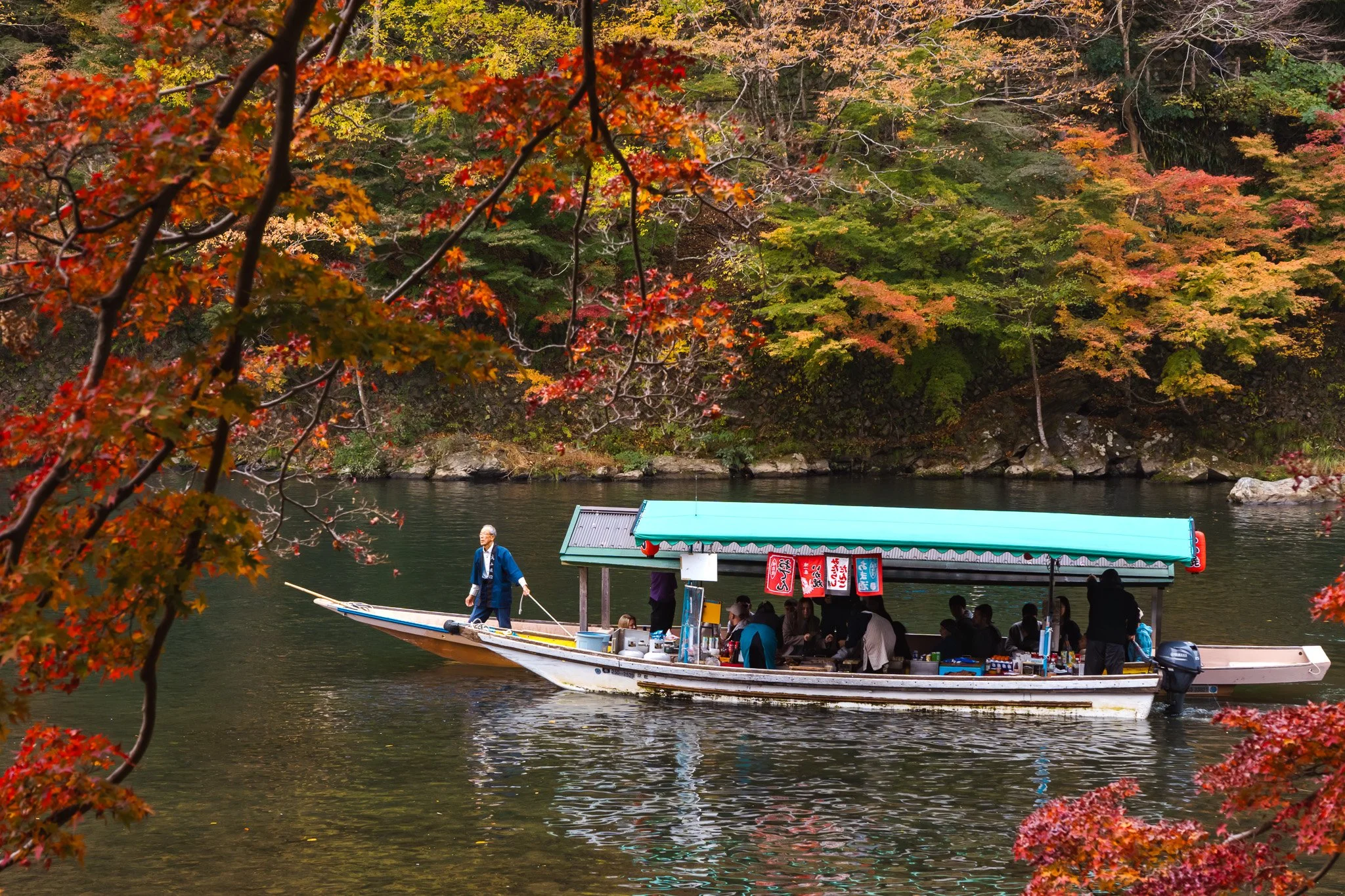 A boat with a green canopy floating on the Arashiyama river during autumn, with red and orange foliage in the background in Kyoto.