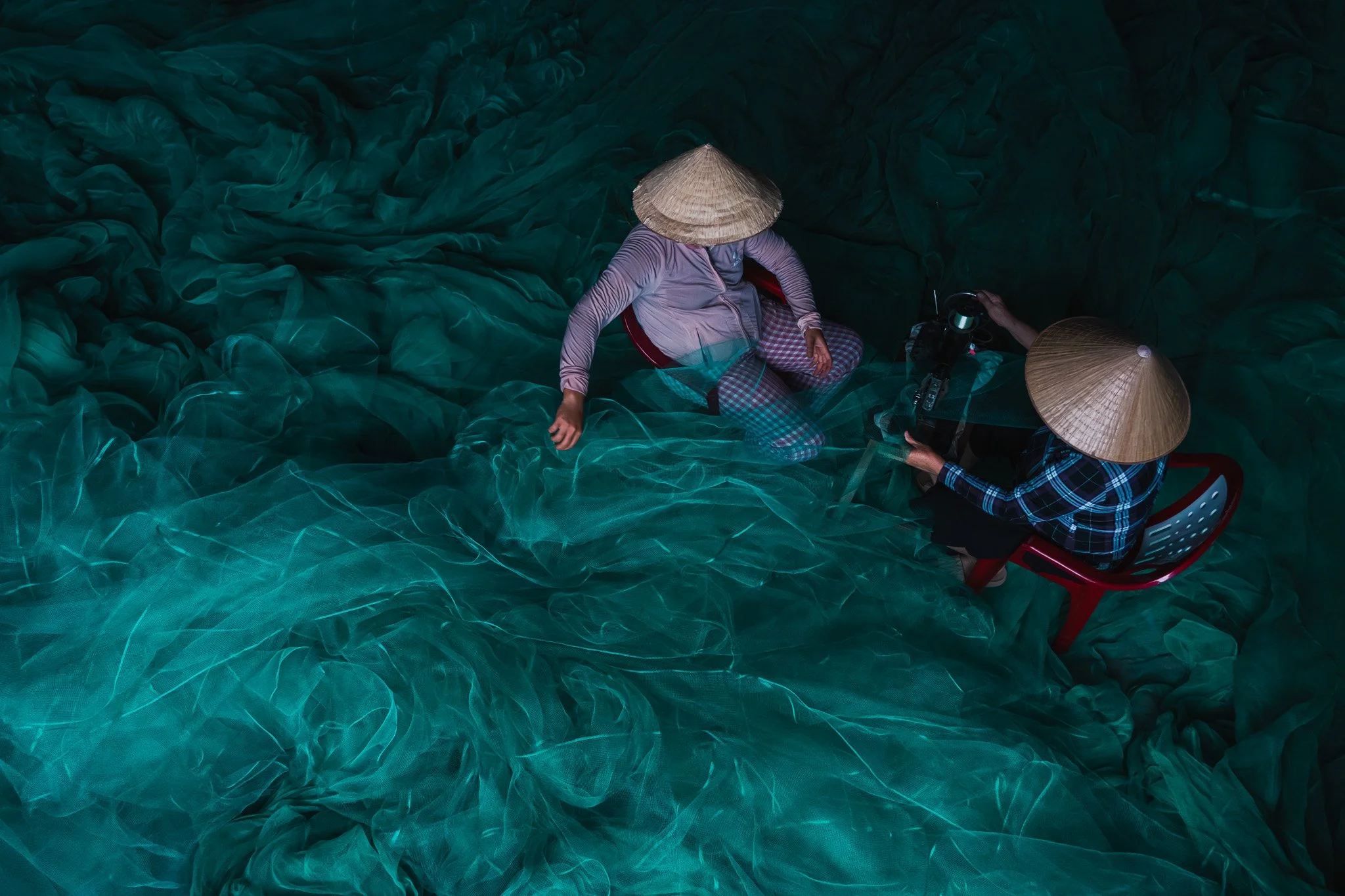 Two people wearing traditional Asian conical hats are sitting on a large green net, helping each other sow the net.