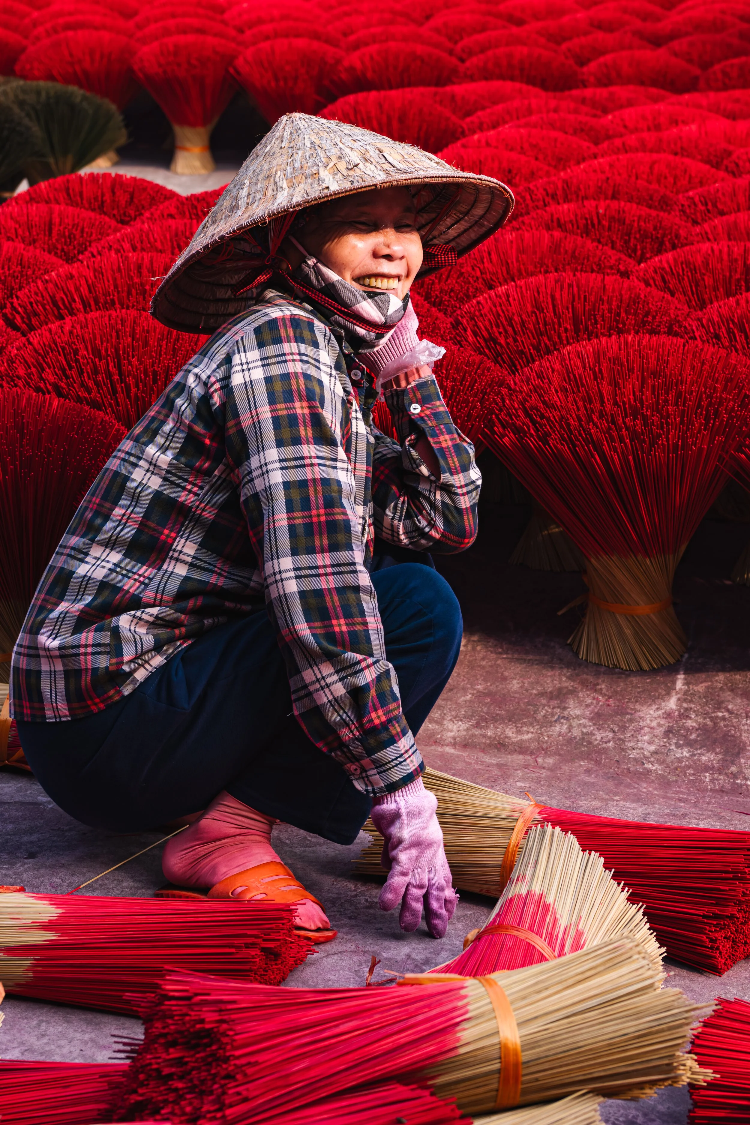 A smiling Vietnamese woman wearing a plaid shirt, traditional conical hat, and pink slippers, crouching among bundles of red incense sticks.