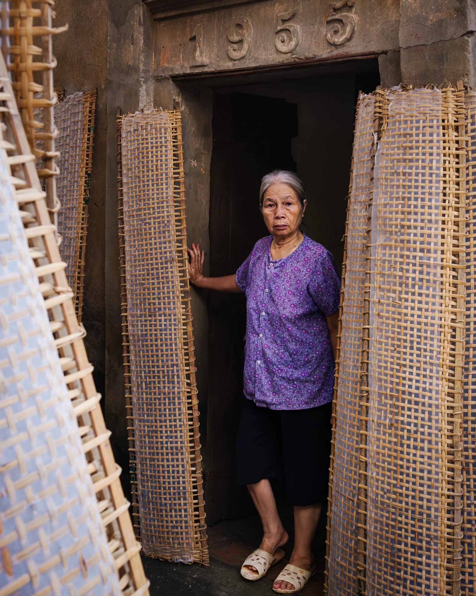 An elderly woman in a purple floral shirt and black skirt stands in a doorway, holding a woven bamboo curtain. The doorway has a concrete frame with the numbers 1955 on top, and she is barefoot wearing beige slippers.