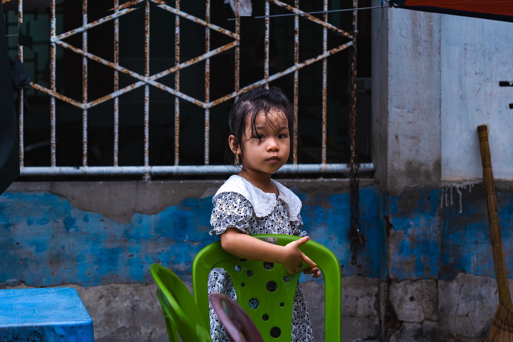 A young girl standing outdoors with a green plastic chair, wearing a black and white dress with lace details and holding onto the back of the chair, in front of a weathered wall with patches of blue paint. There is a rusty metal gate and a broom lean