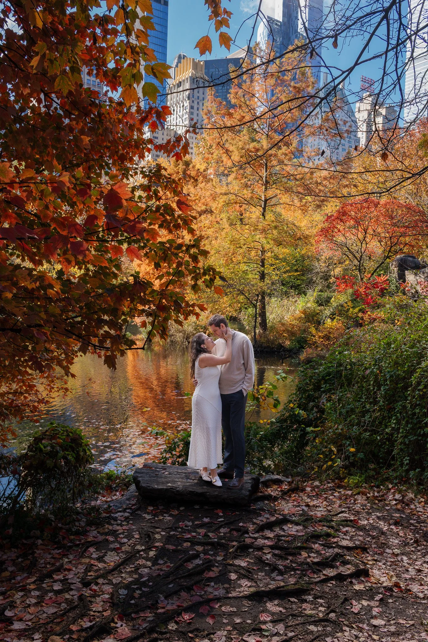 A couple stands on a log by a pond surrounded by autumn trees in Central Park, with tall buildings in the background.