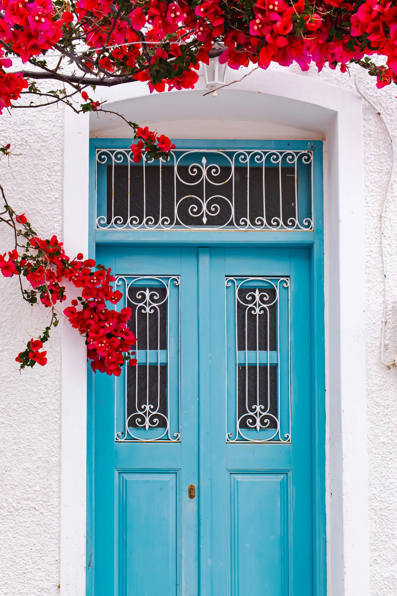Bright blue double door with decorative white ironwork, surrounded by white textured walls and vibrant pink bougainvillea flowers at the top left corner.