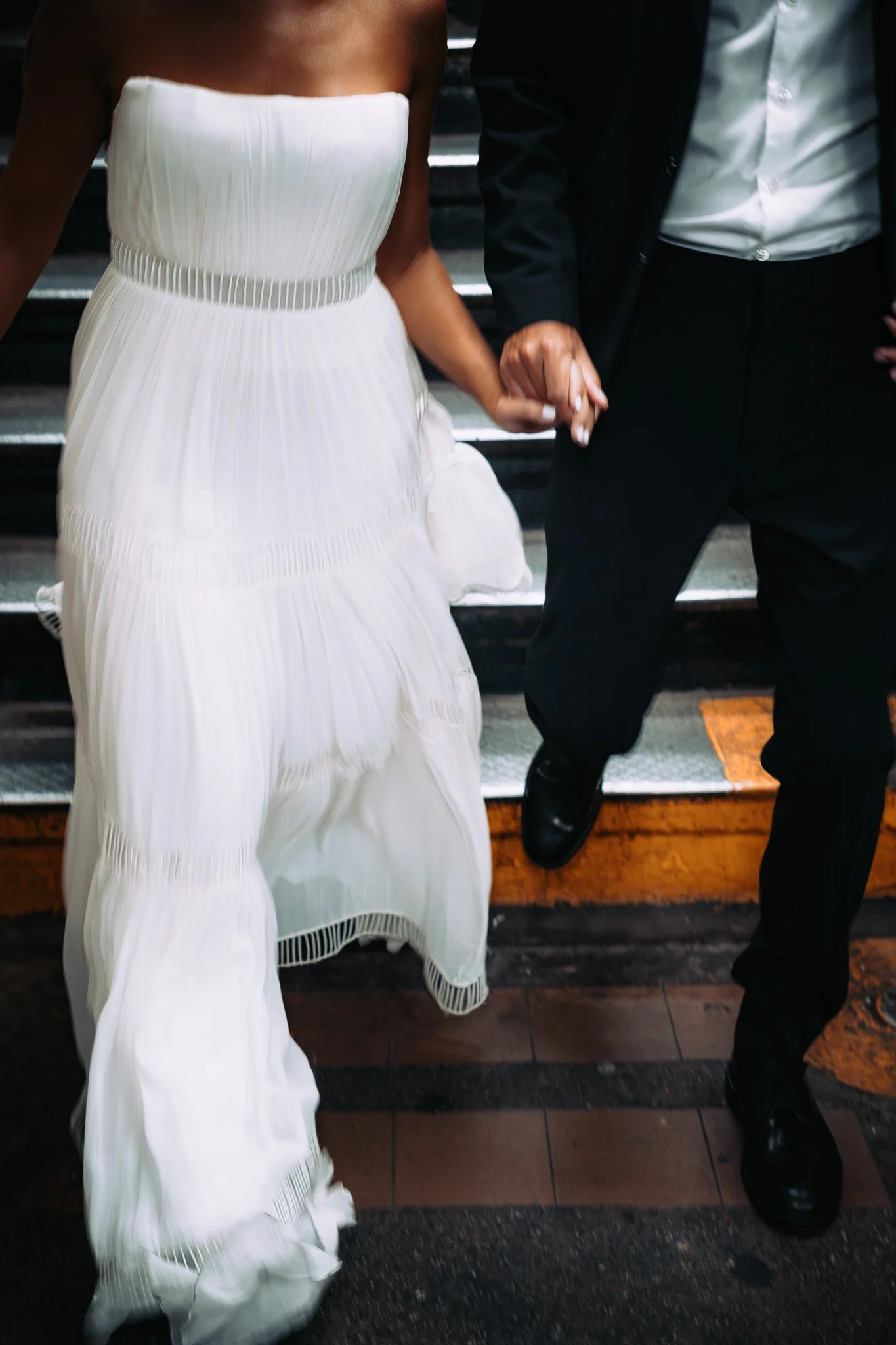 A couple holding hands, the woman in a white dress and the man in a black suit, running down the NYC subway stairs.