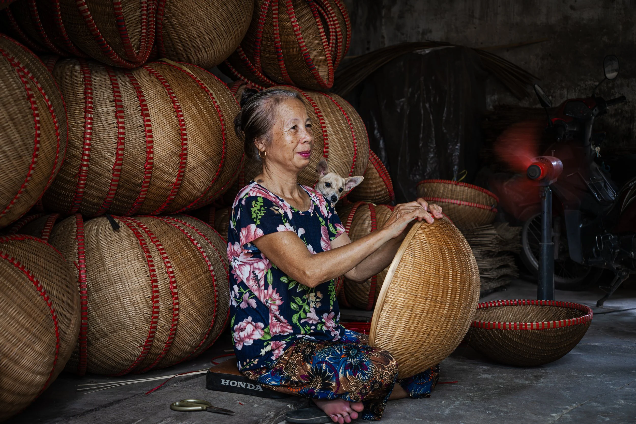 An elderly woman with gray hair and wearing a floral shirt, sitting cross-legged on the ground in front of stacked woven baskets, holding one basket and a small dog on her shoulder. There are more baskets and a motorcycle in the background.