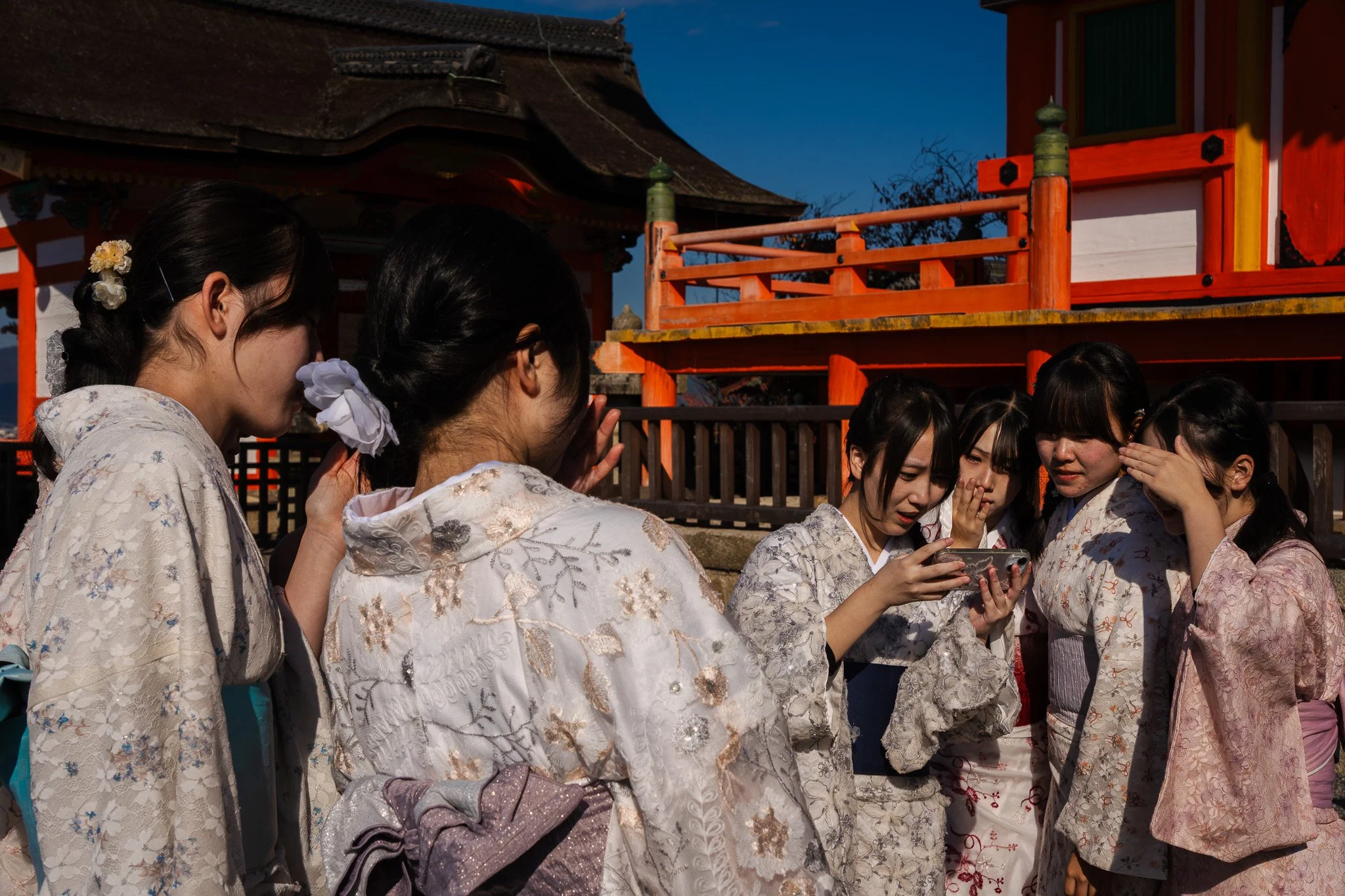 Group of women wearing traditional kimono garments, some bowing, some looking at a smartphone, outside Kiyomizu-dera Temple, a Japanese shrine, during daylight.