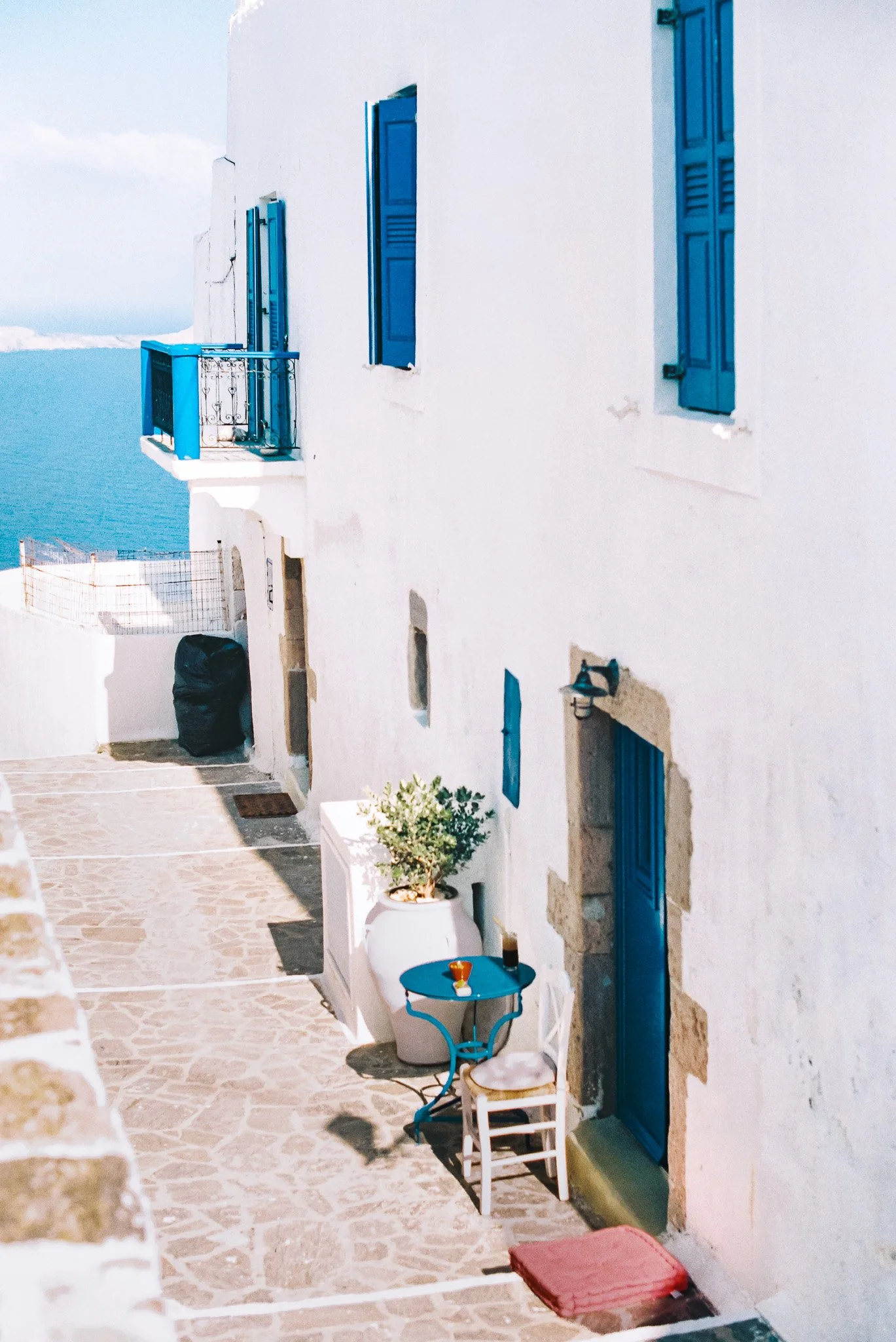 White Mediterranean building with blue shutters and door, overlooking the ocean, with outdoor table and chairs, potted plant, and decorative elements.
