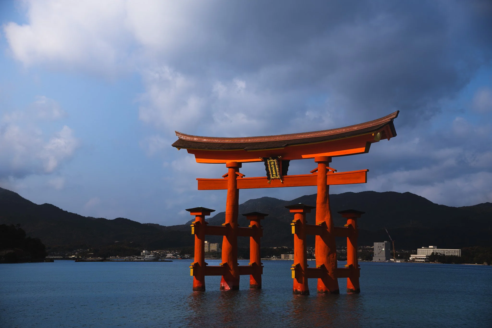 Traditional Japanese torii gate in Miyajima Island, standing in water with mountains and cloudy sky in background.