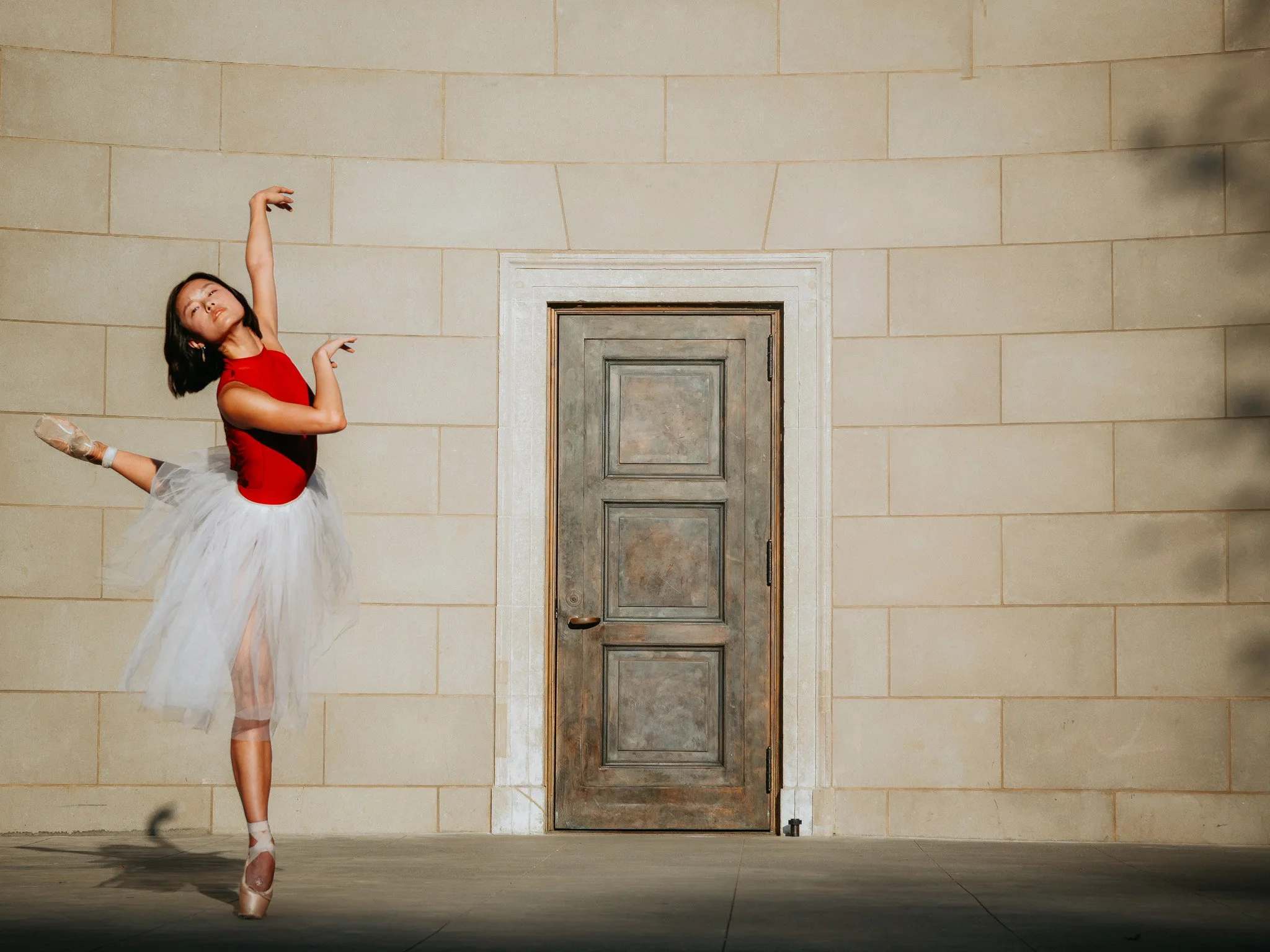 A ballerina in a red leotard and white tutu performing a balanced pose on pointe at Naumburg Bandshell with a closed wooden door in Central Park.