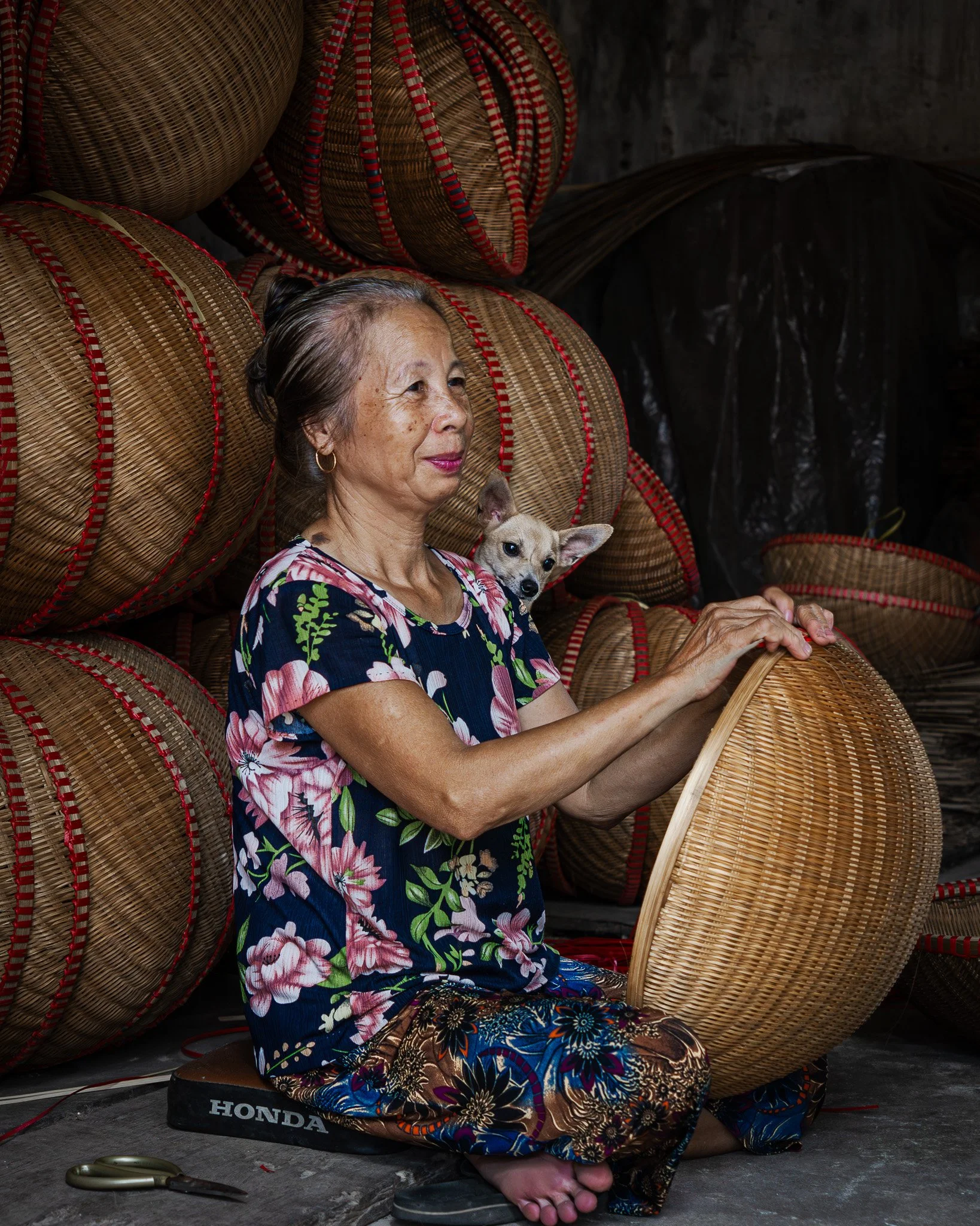 An elderly woman sitting on the floor with a small dog on her shoulder, surrounded by bamboo baskets, in a workshop area.
