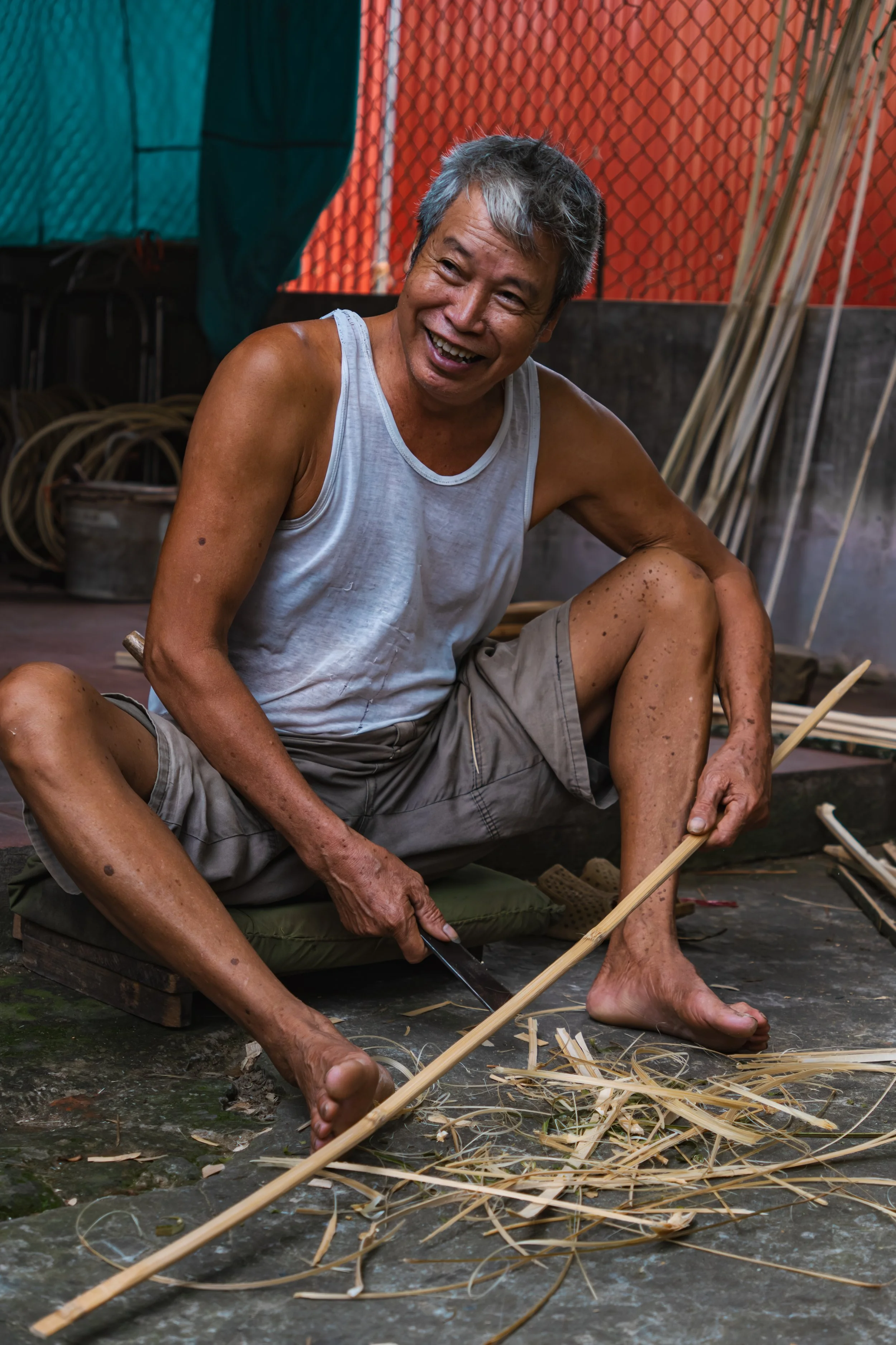 An older man with gray hair, wearing a white tank top and shorts, is sitting cross-legged on a cushion, smiling while carving bamboo with a knife, making a bamboo basket.