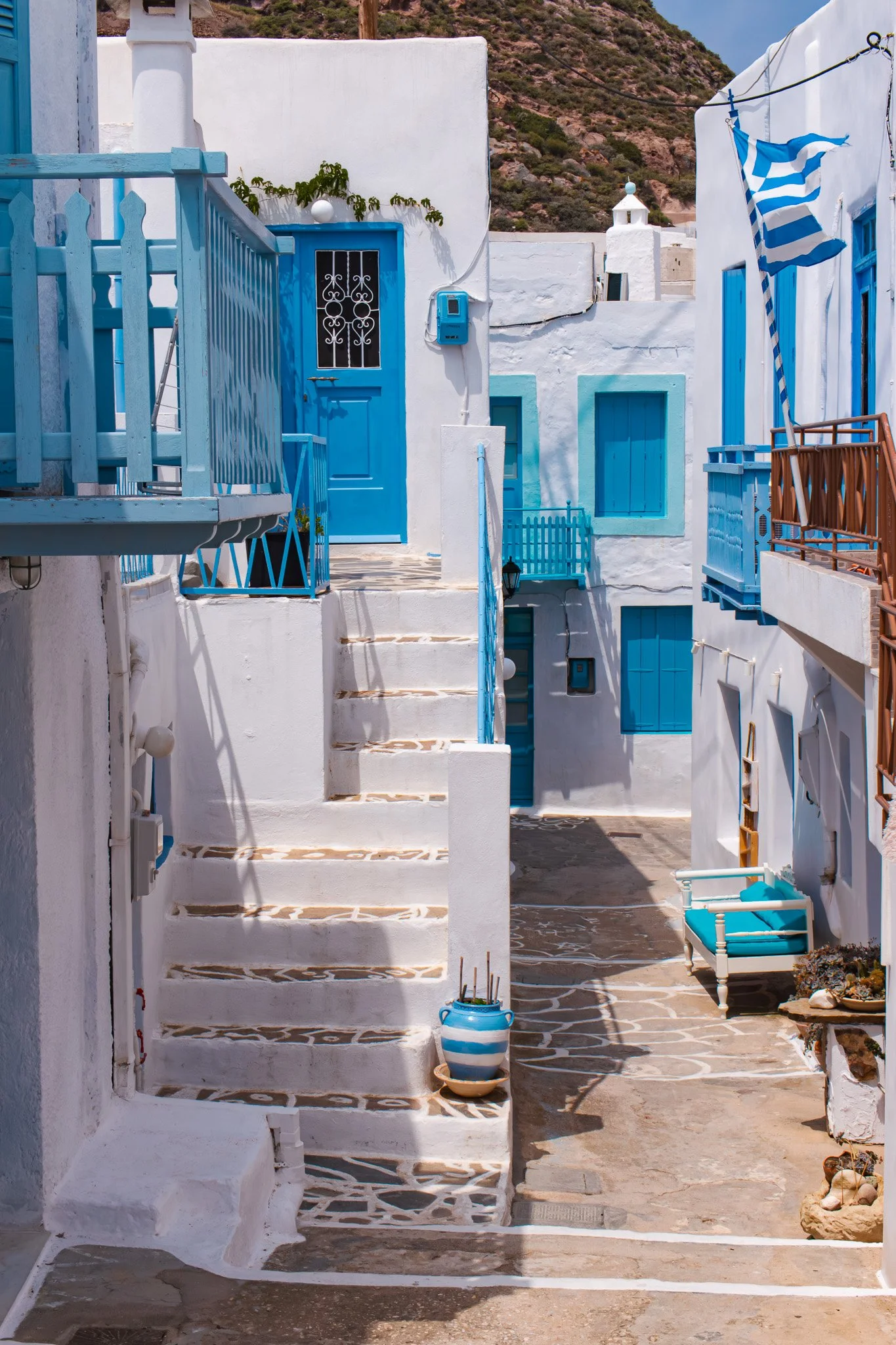 A narrow alley in a Greek village with white walls and blue accents, including doors, windows, and railings, with stairs leading to a door, and a blue-and-white striped flag hanging from a balcony.