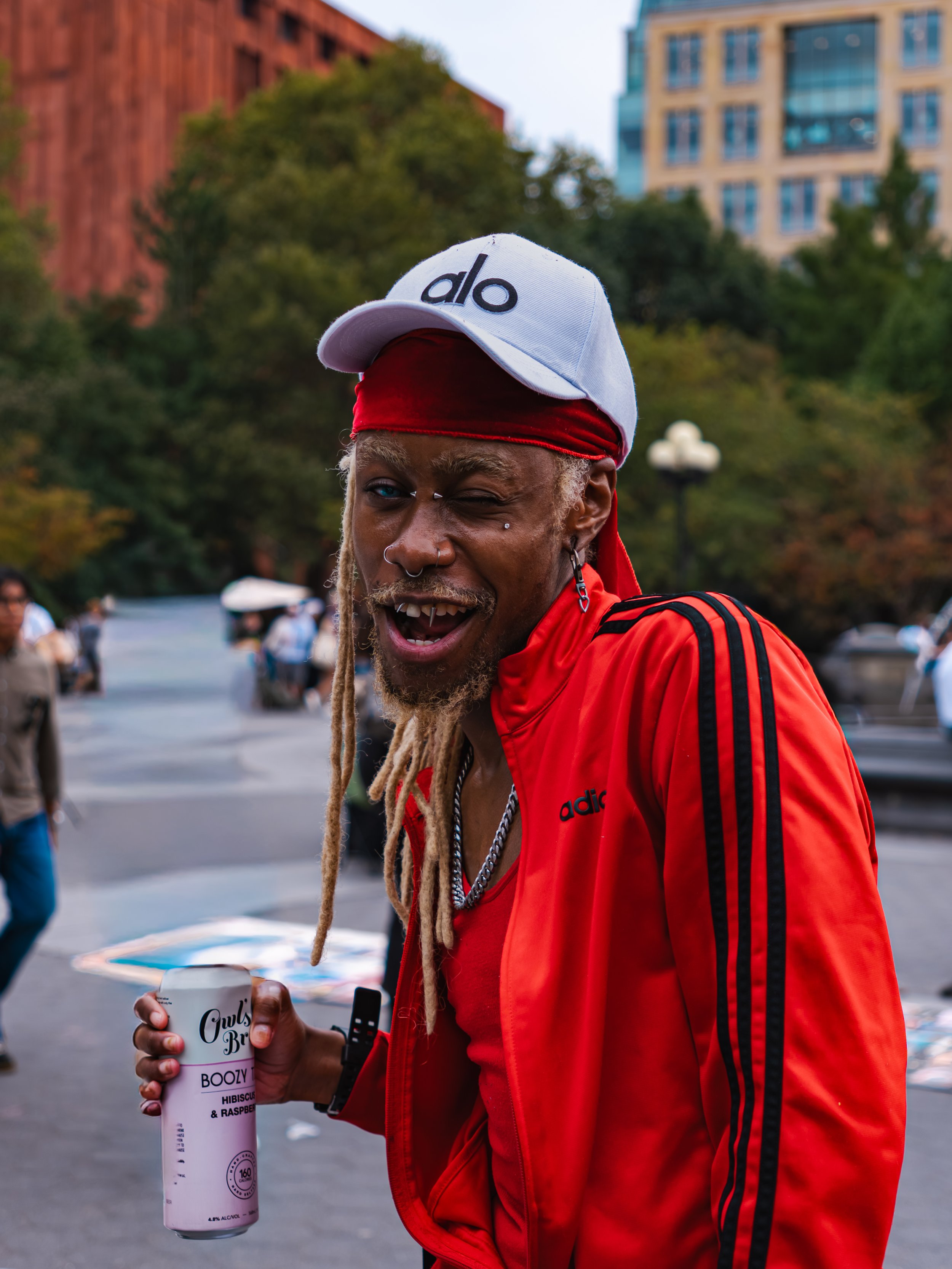 A man with dreadlocks and facial jewelry wears a red adidas jacket, a white 'Alo' cap, and a red bandana, holding a can of drink while smiling at the camera in Washington Square Park, with trees and people in the background.