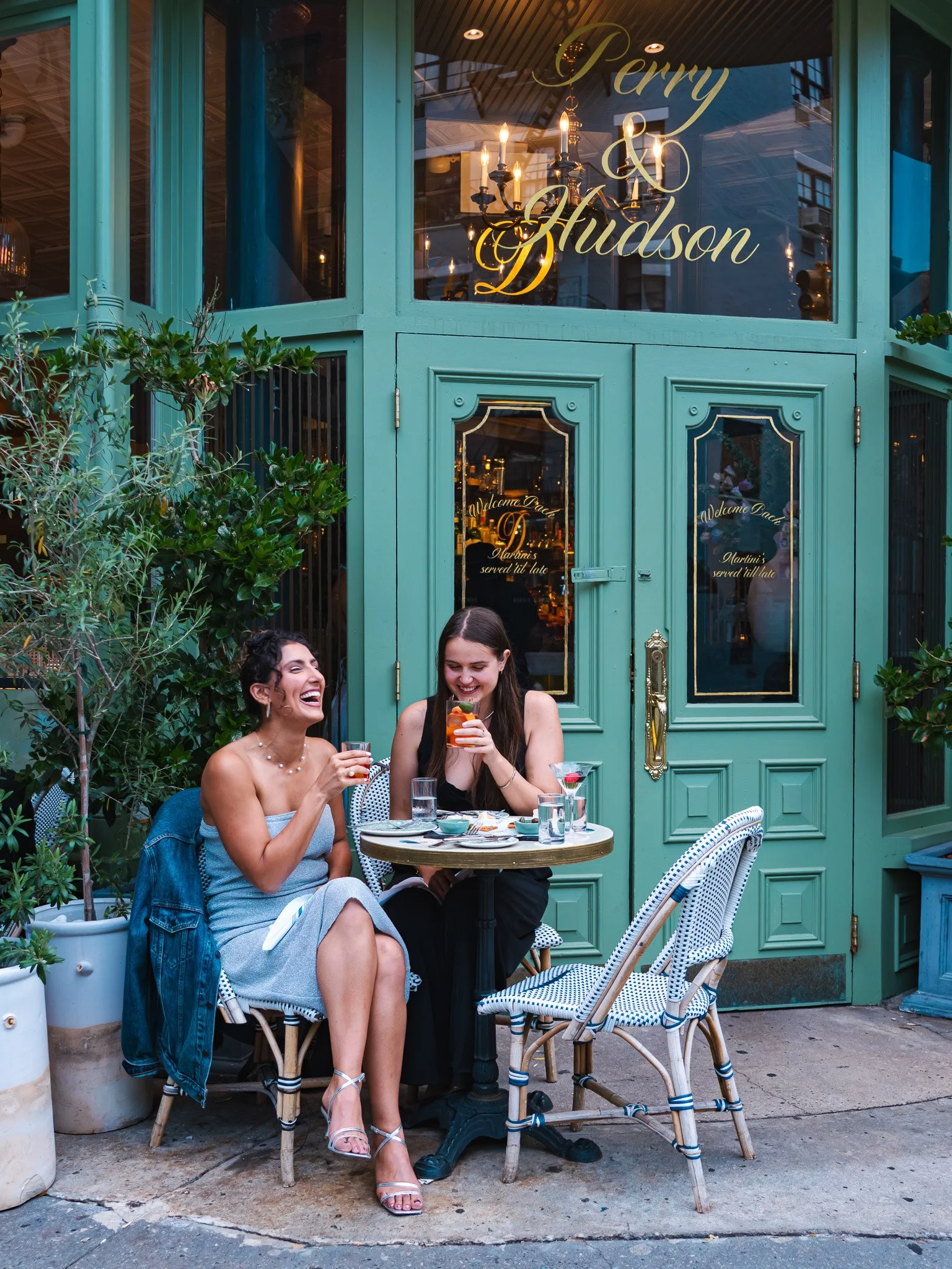 Two women sitting outside Dante in the West Village, enjoying drinks and laughter. The women are seated at a small round table with various drinks and food plates, in front of a green entrance with large windows and decorative text.