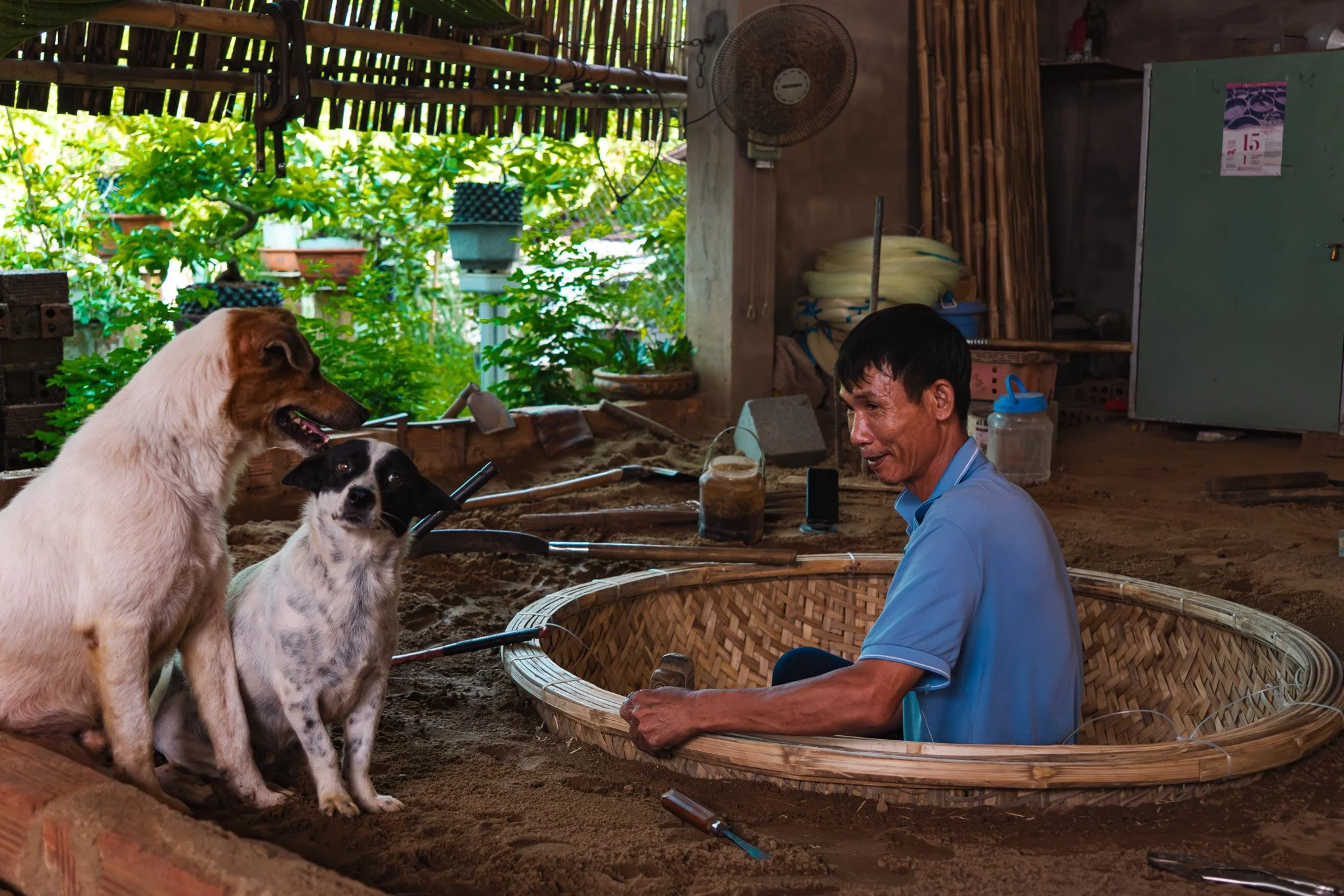 A man working with two dogs in a rustic indoor space with a green garden outside. The man is sitting in a round woven boat, holding a tool, with one dog sitting beside him and another standing nearby.