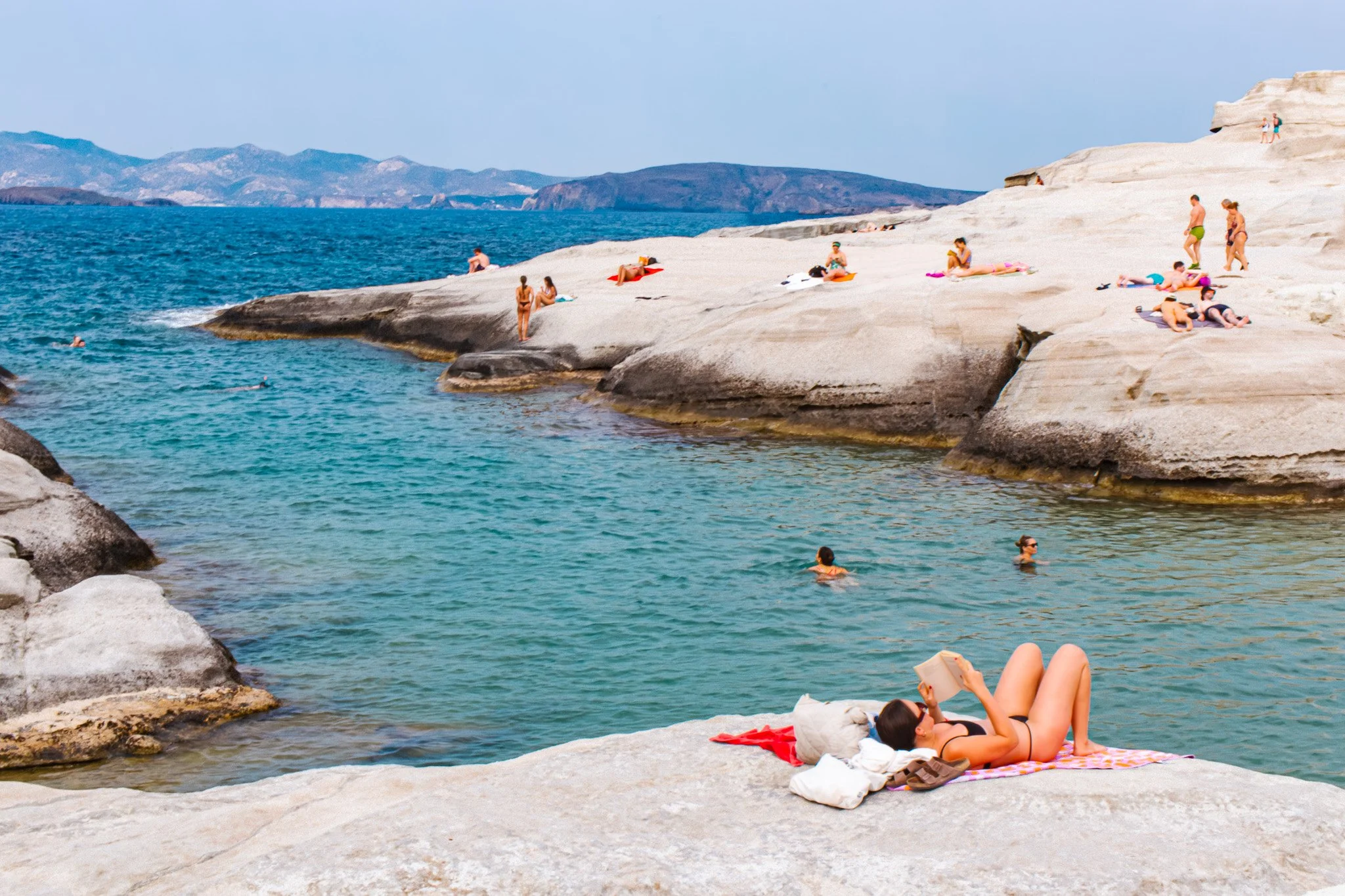 People relaxing on rocks and swimming in the water at Sarakiniko beach in Milos with mountains in the background.