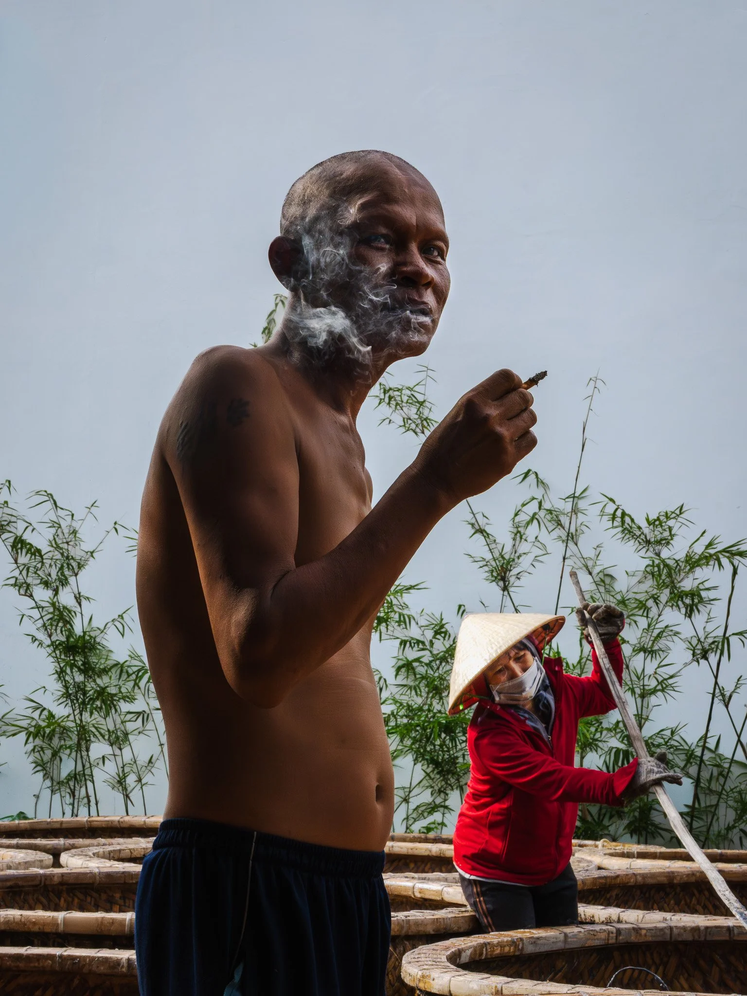 A man with a shaved head and bare torso is smoking a cigarette, with smoke around his face, standing outdoors. In the background, a person in a red jacket, face mask, and traditional conical hat is working with a long tool near bamboo baskets, with t