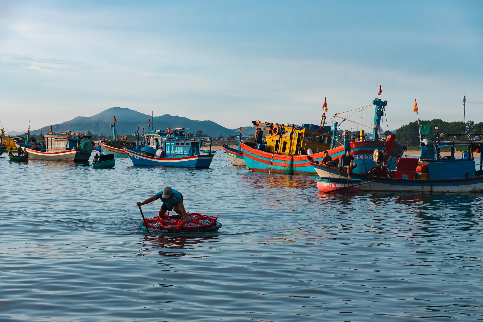 Colorful fishing boats docked by the waterfront with mountains in the background, and a person paddling a small raft in the water in the foreground.