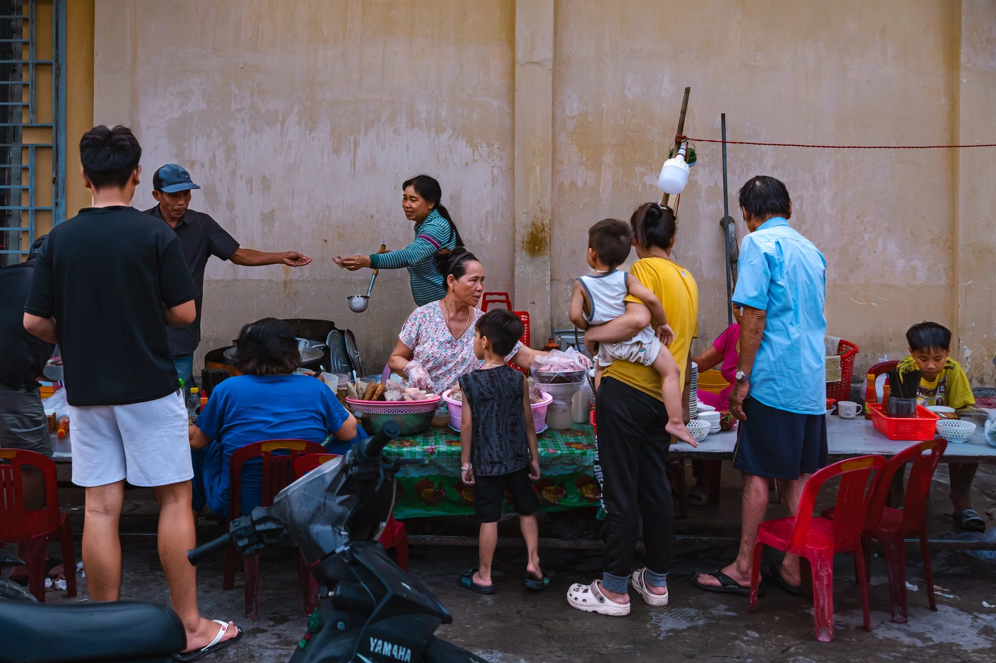 A group of people gathered around a table at an outdoor food market. The scene shows various individuals serving and receiving food, including children and adults, with some seated and others standing. The setting appears to be in a narrow alley with