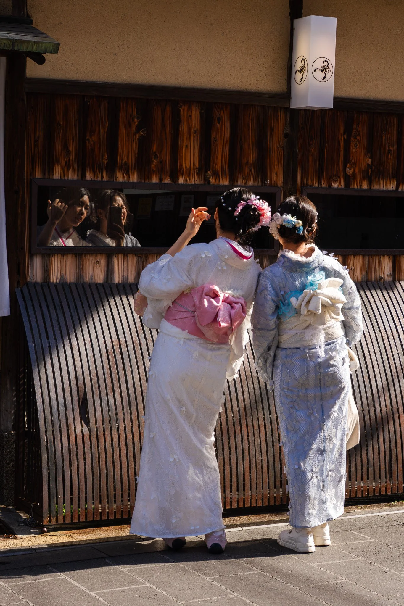 Two women wearing traditional Japanese kimonos with floral hair adornments, standing outside a building, looking into a window with their reflections visible in Kyoto.