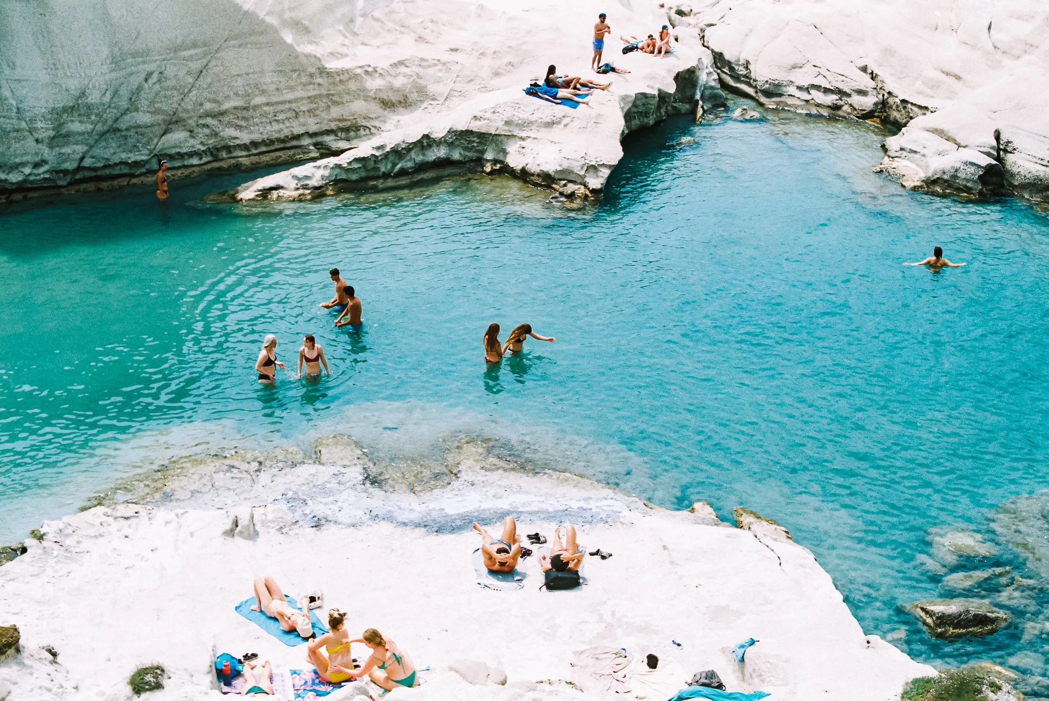 People swimming, lounging, and sunbathing at a rocky turquoise lake and white rocks of Sarakiniko beach in Milos, Greece. 