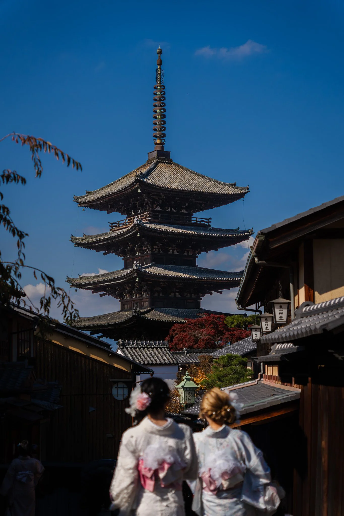 Hōkan-ji Temple (Yasaka Pagoda), a traditional Japanese five-story pagoda in a temple complex, with two women dressed in kimonos walking in the foreground, under a clear blue sky.