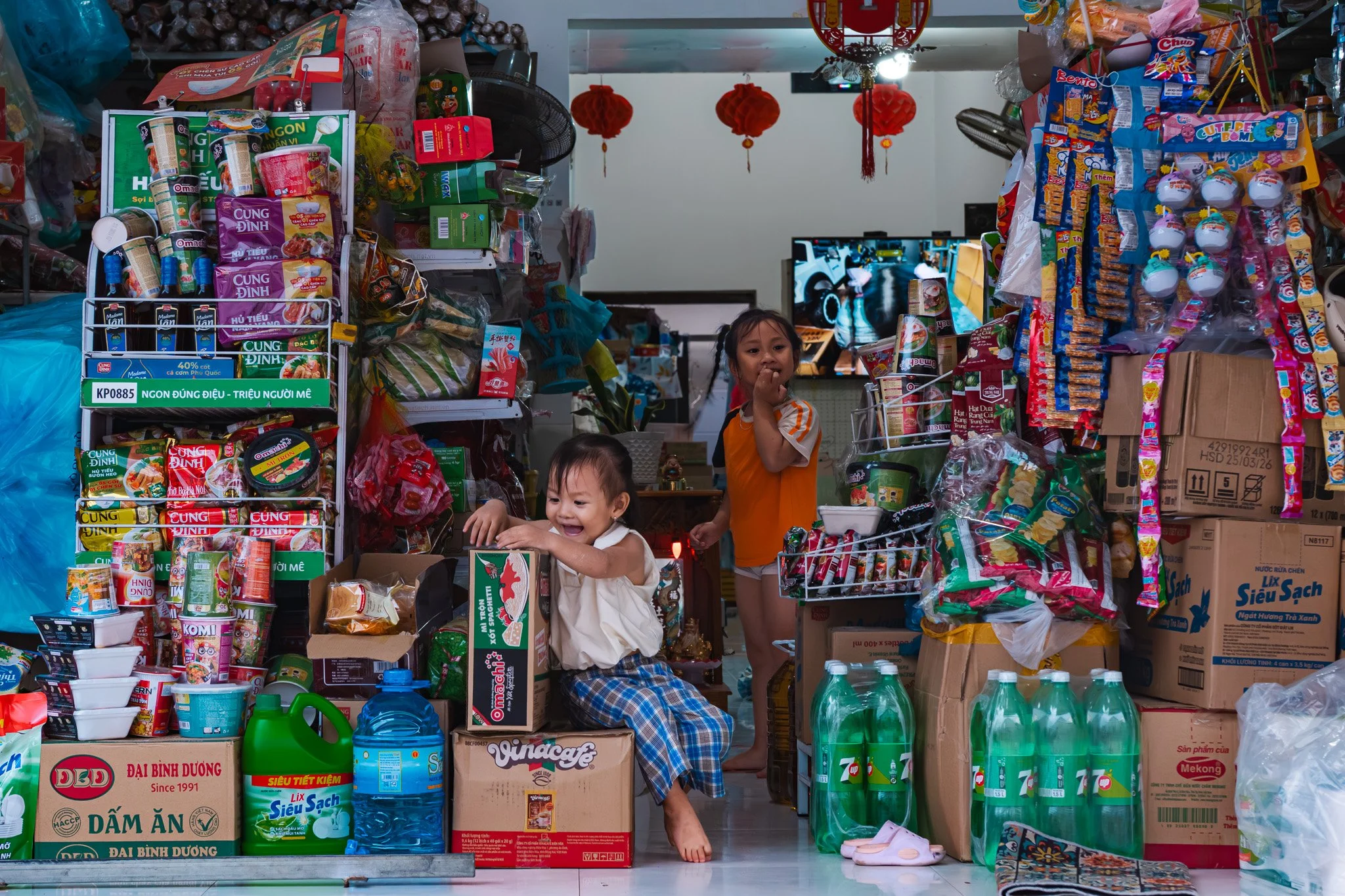 Two children, a girl in an orange shirt and a younger girl in plaid pants and a cream shirt, play and smile inside a cluttered grocery store filled with snacks, drinks, and household items.