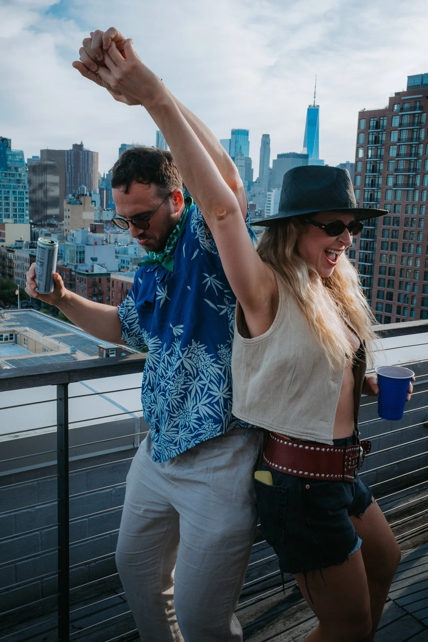 A man and woman dancing on a rooftop with a city skyline in the background, holding drinks.