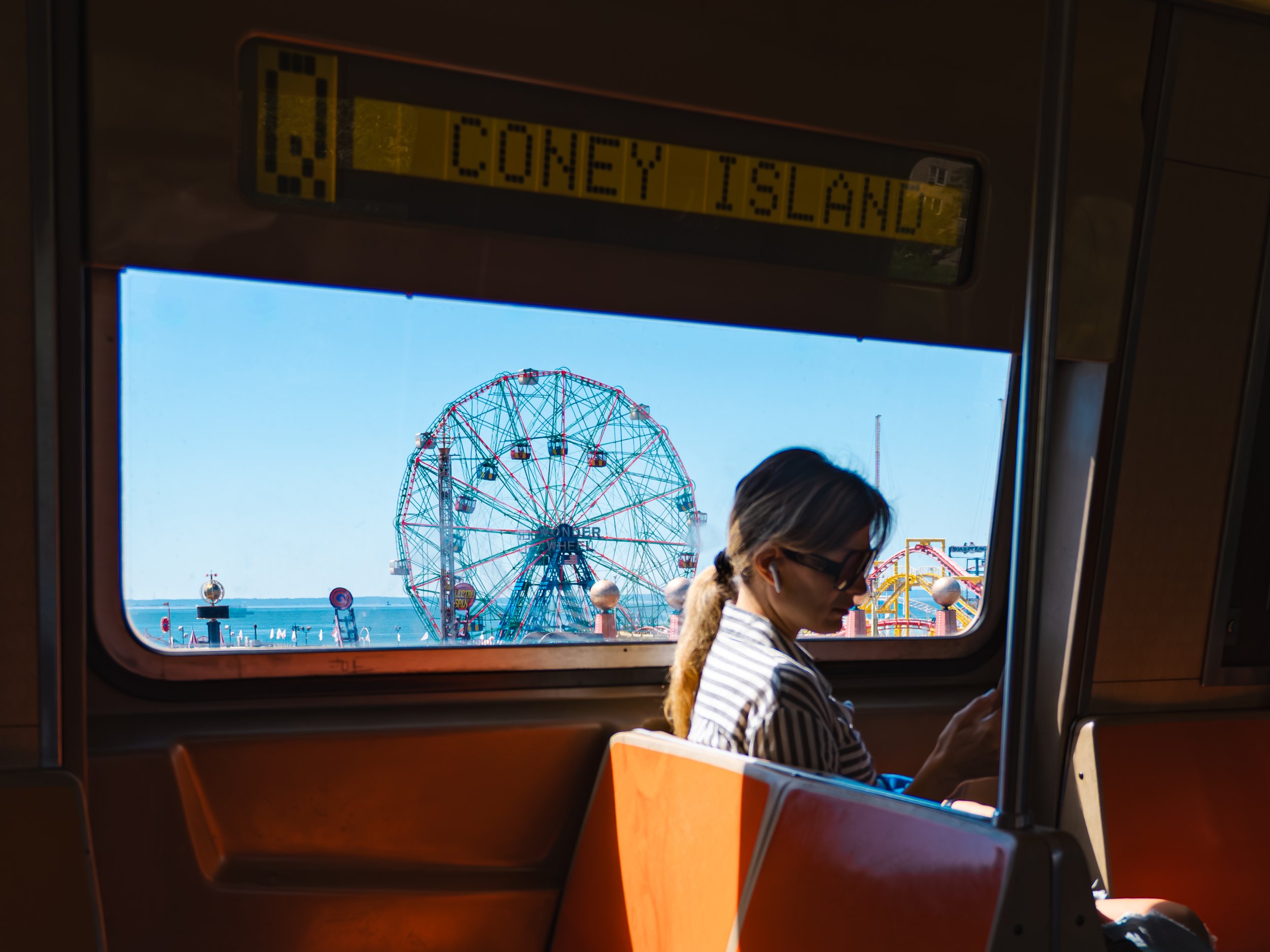 A woman sitting on the Q train in New York, looking at her phone, with a view of a Ferris wheel, amusement park rides, and the ocean through the window, indicating a seaside amusement park in Coney Island.