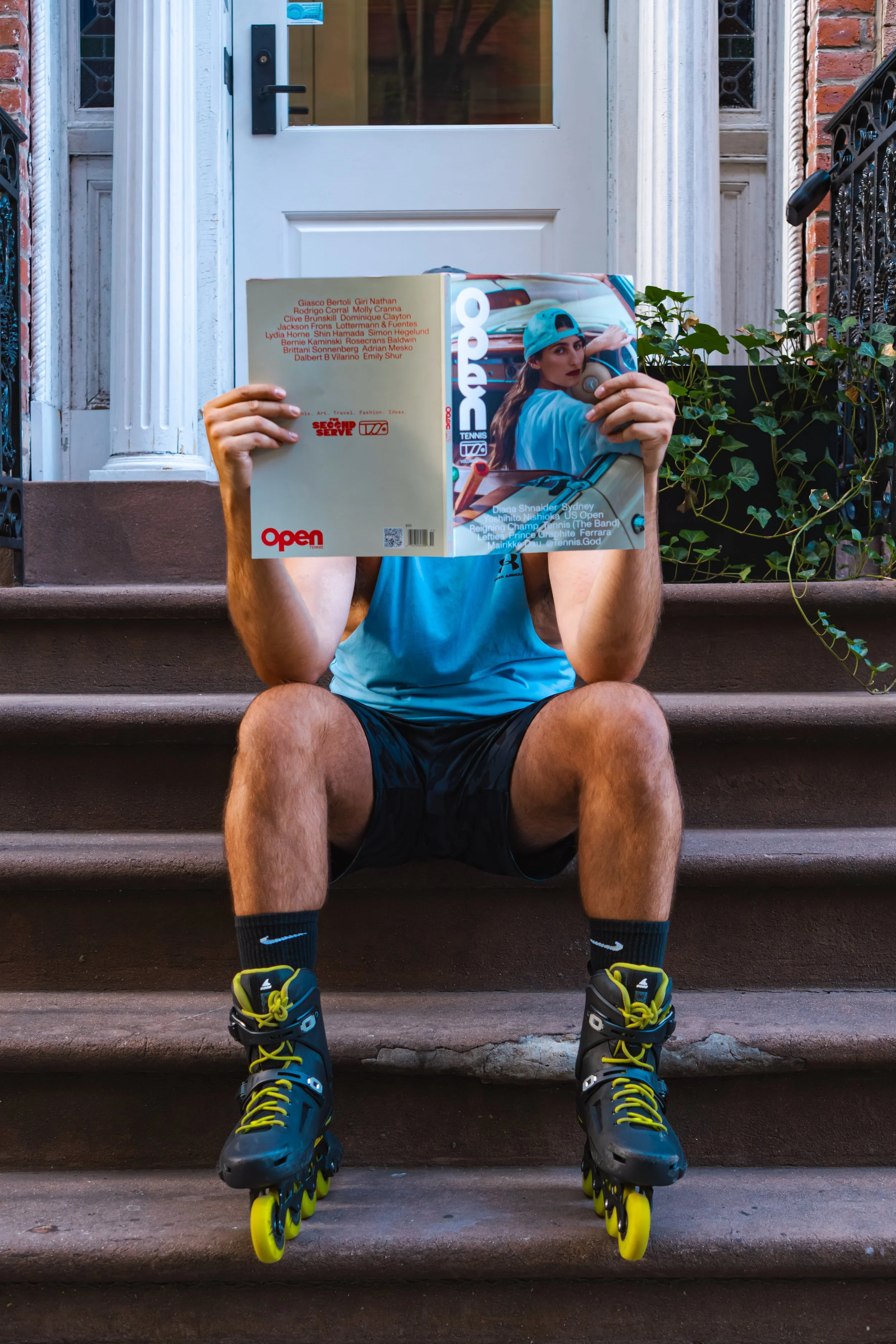 Person sitting on brown stairs wearing black rollerblades, black socks with blue Nike logo, blue sports shorts, and a light blue athletic shirt, reading a magazine that covers their face, with the magazine featuring a woman in a blue cap and sportswe