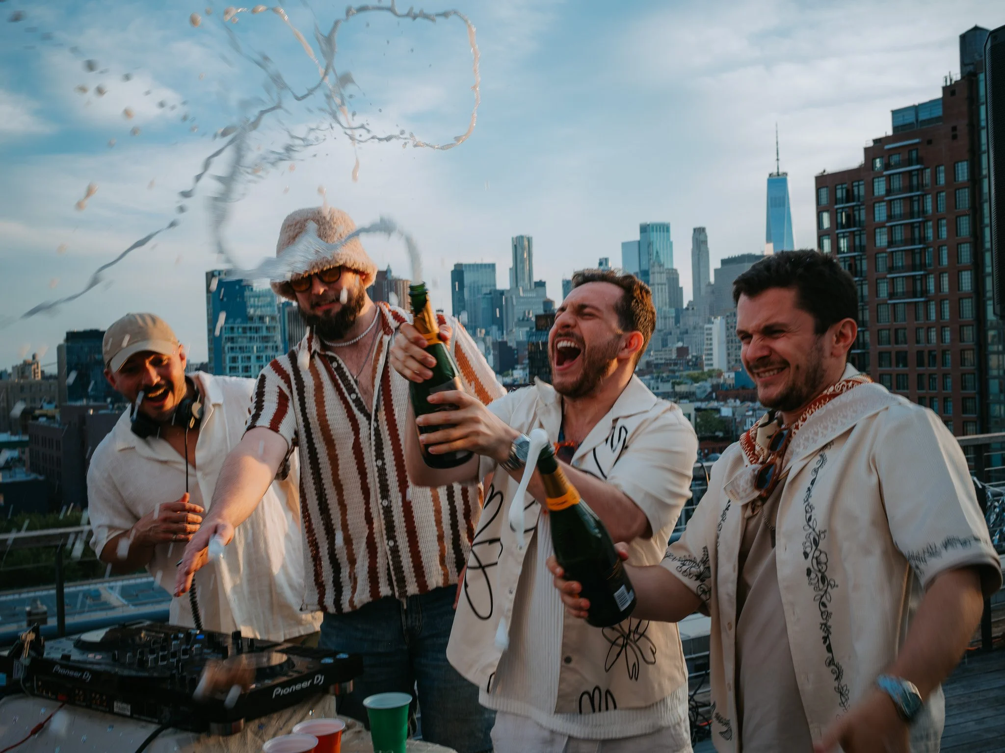 Four men celebrating on a NYC rooftop with city skyline in the background, holding champagne bottles, with one spraying champagne.