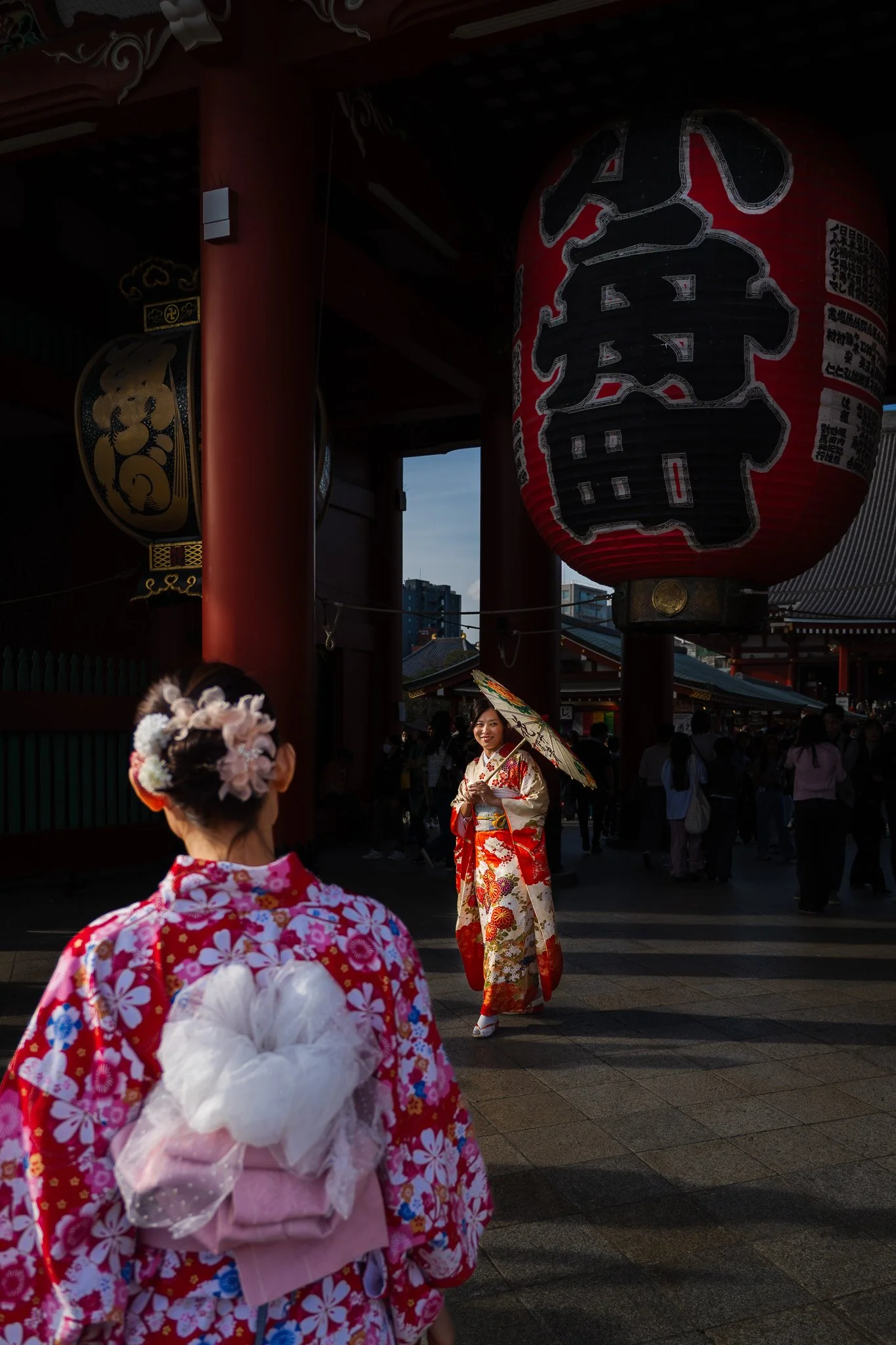 Two women wearing traditional Japanese kimonos, one holding an umbrella, in front of a large red paper lantern Senso-ji temple during the day.