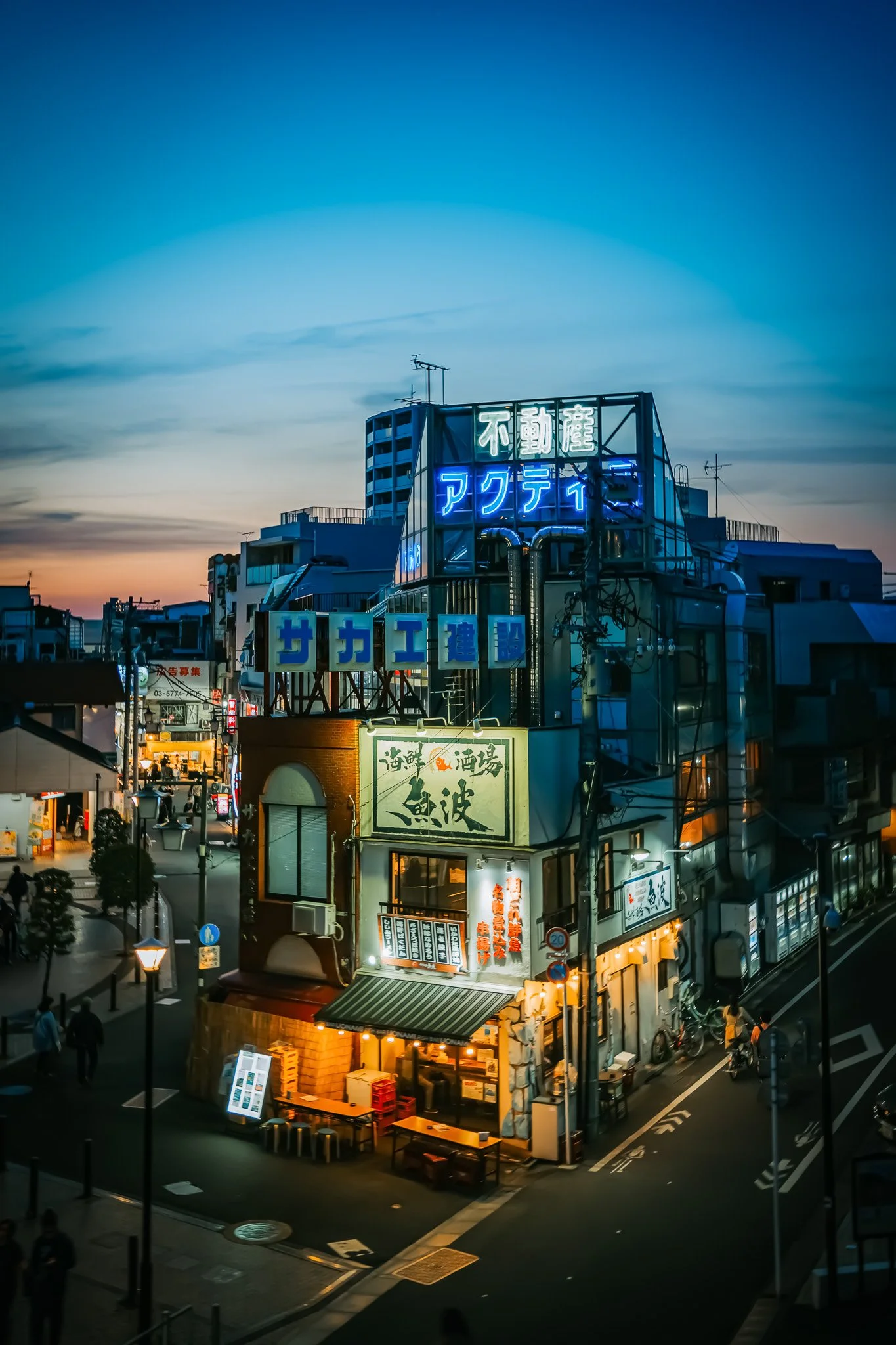 Night view of a city street with illuminated signs in Japanese, storefronts, and a few pedestrians.