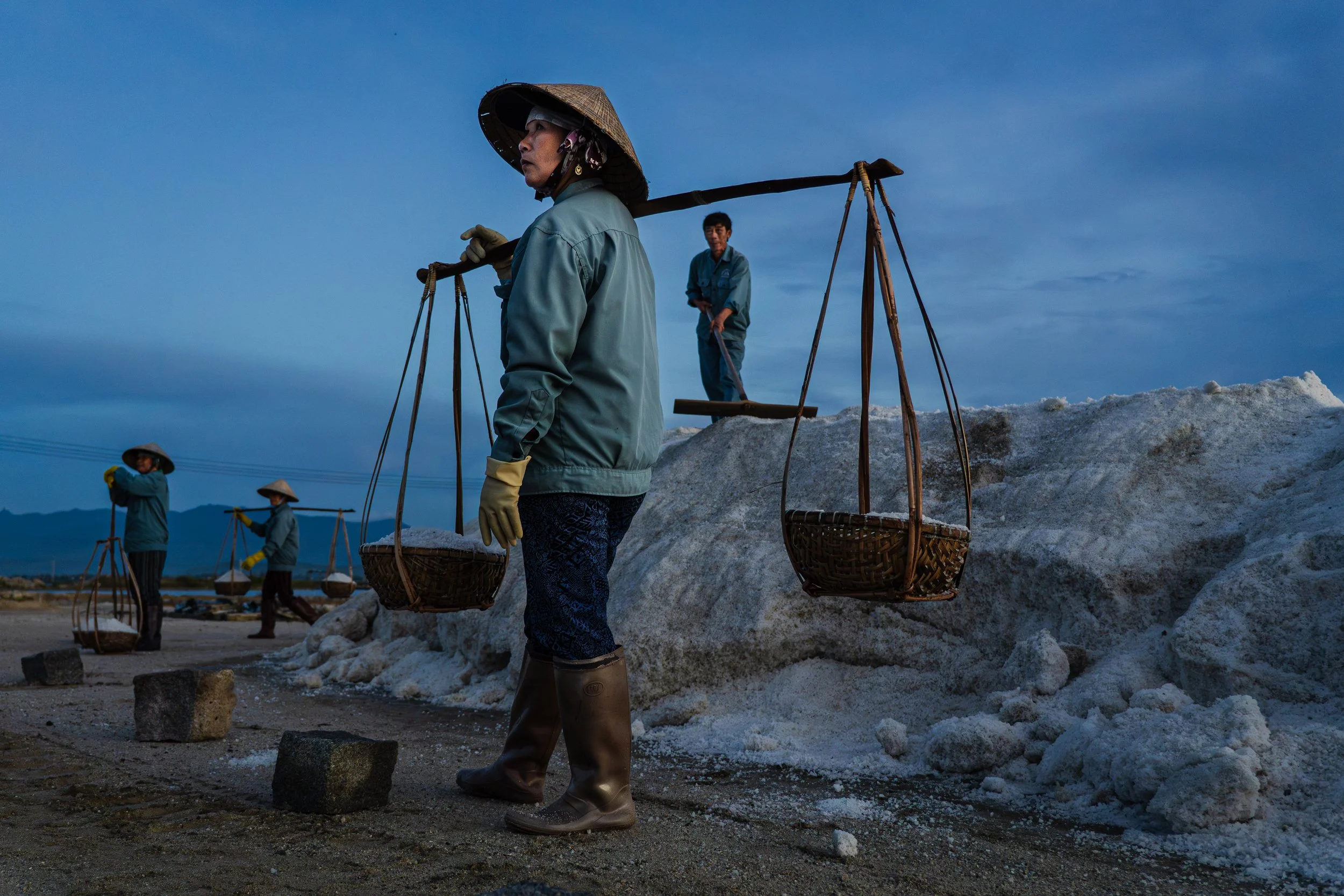 Vietnamese workers standing by large pile of salt at an outdoor salt flat, wearing traditional Asian conical hats and blue uniforms, carrying baskets on shoulder poles.