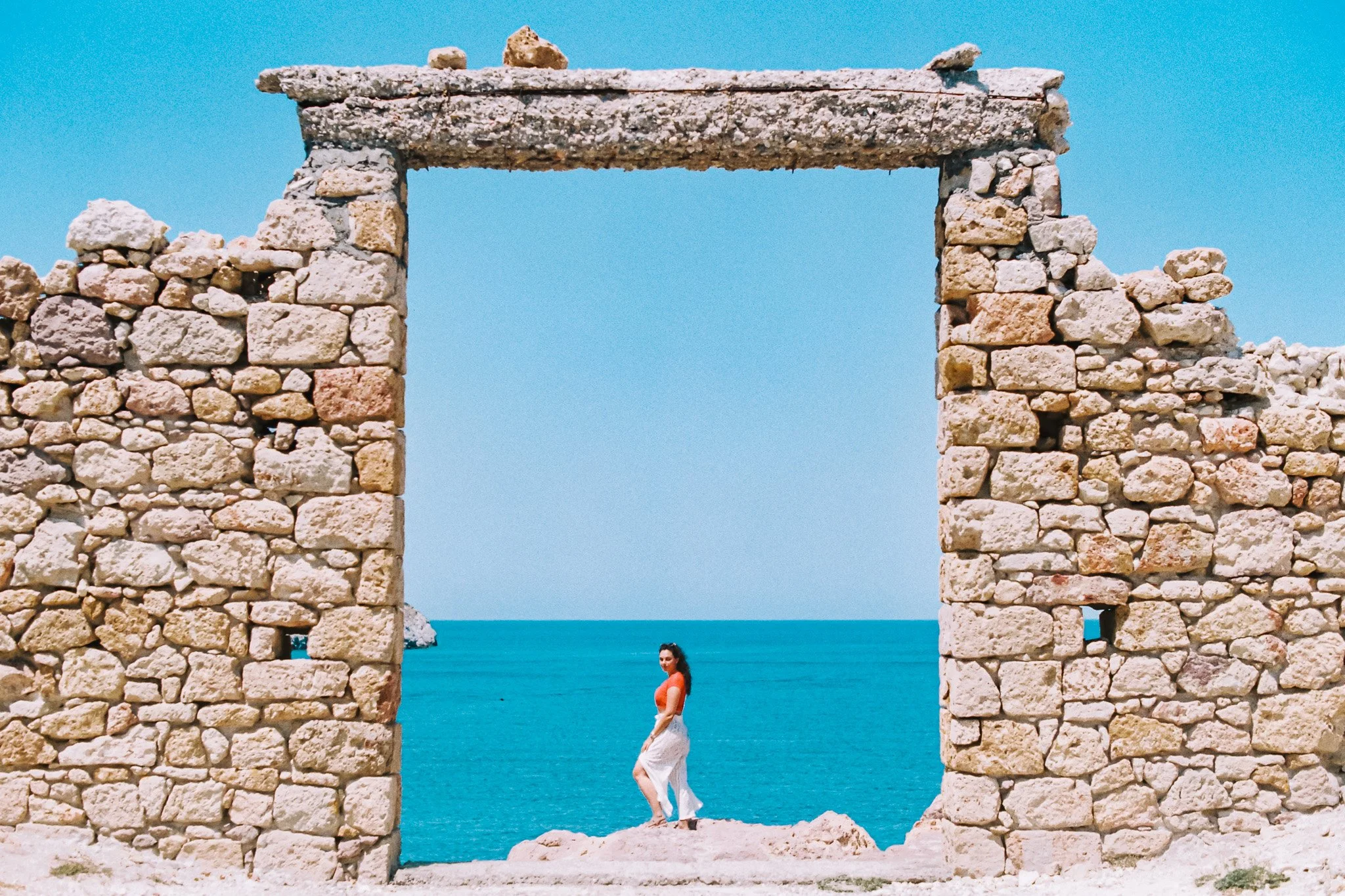 A woman standing on rocks in front of the ocean, framed by an ancient stone wall opening with a bright blue sky in the background.