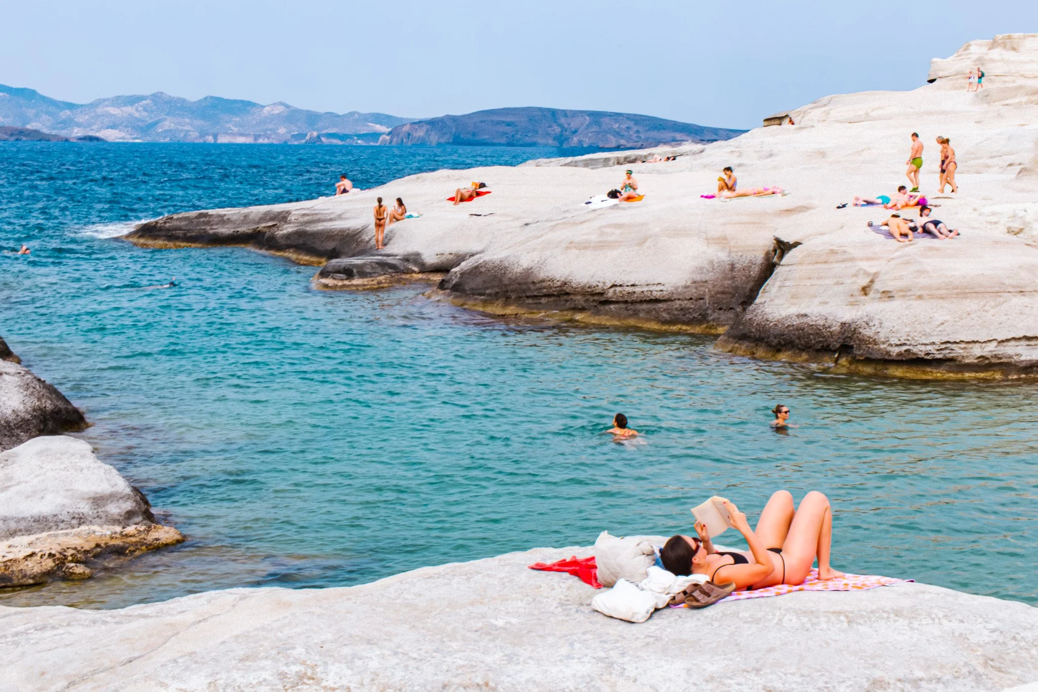 People relaxing and swimming at a rocky shoreline by the ocean in Milos at Sarakiniko beach, with some sunbathing on the rocks and others swimming in the water.