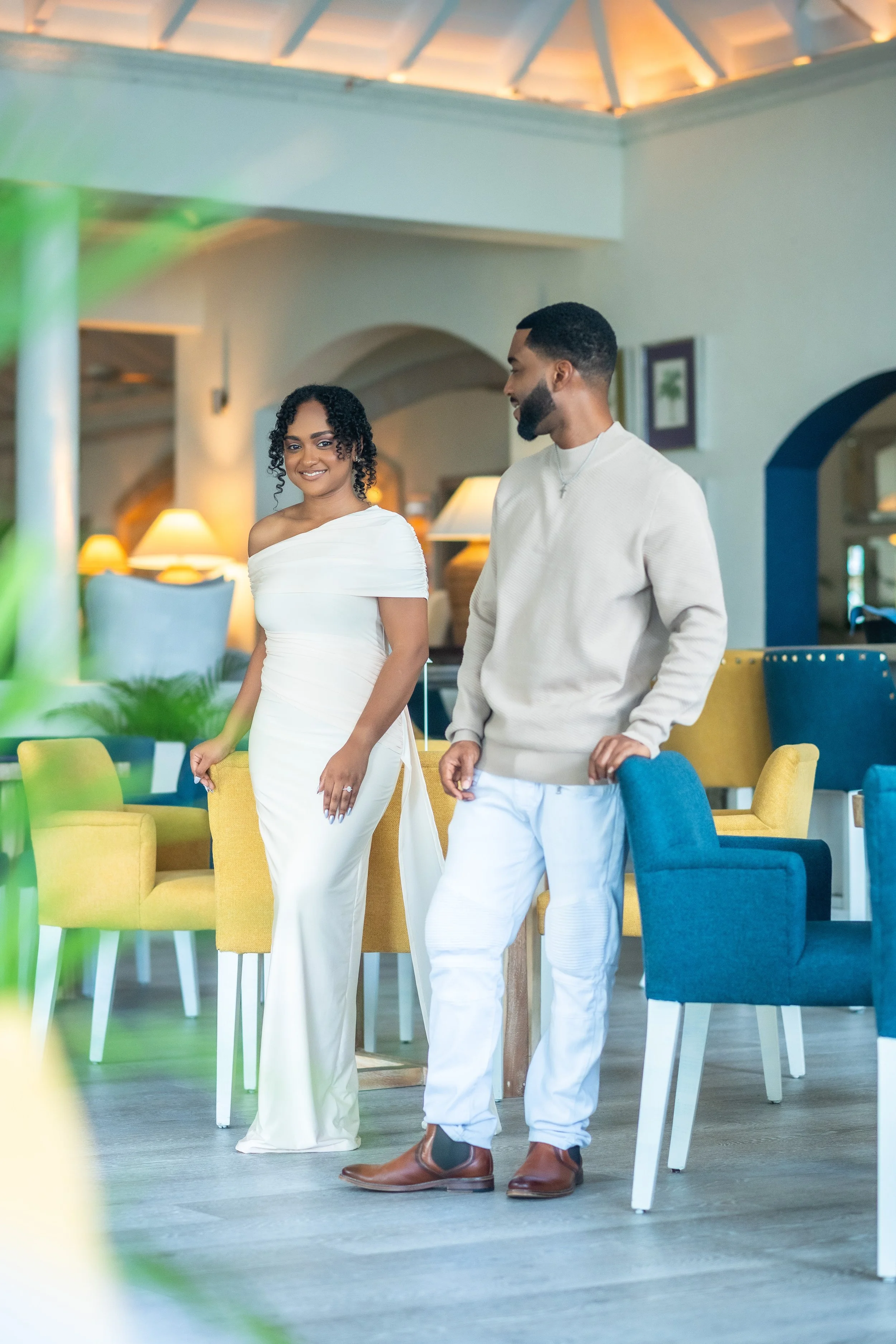 A man and woman inside a well-lit, modern cafe or lounge. The woman is wearing a white, off-the-shoulder gown and smiling, while the man is dressed casually in a beige sweater, white pants, and brown shoes, looking at her.