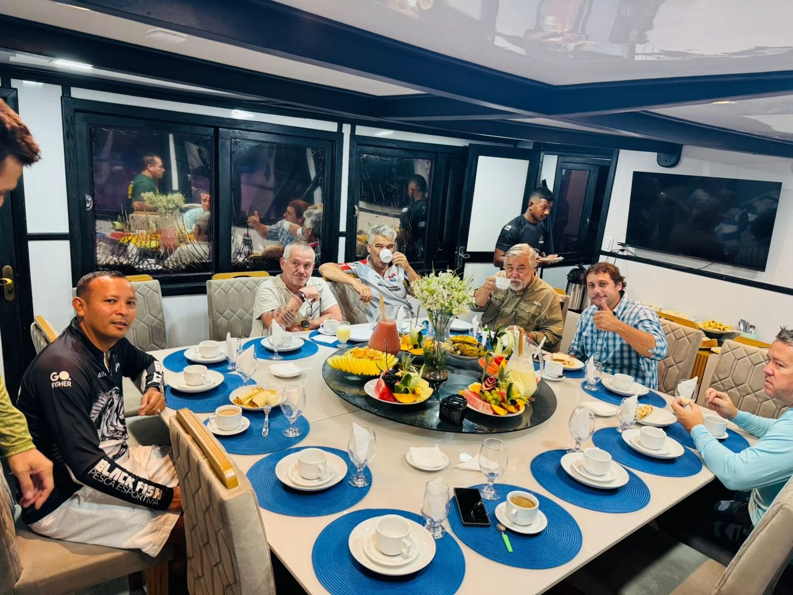 People sitting around a dining table with breakfast foods, drinks, and fruit, in a room with large windows and a TV on the wall.