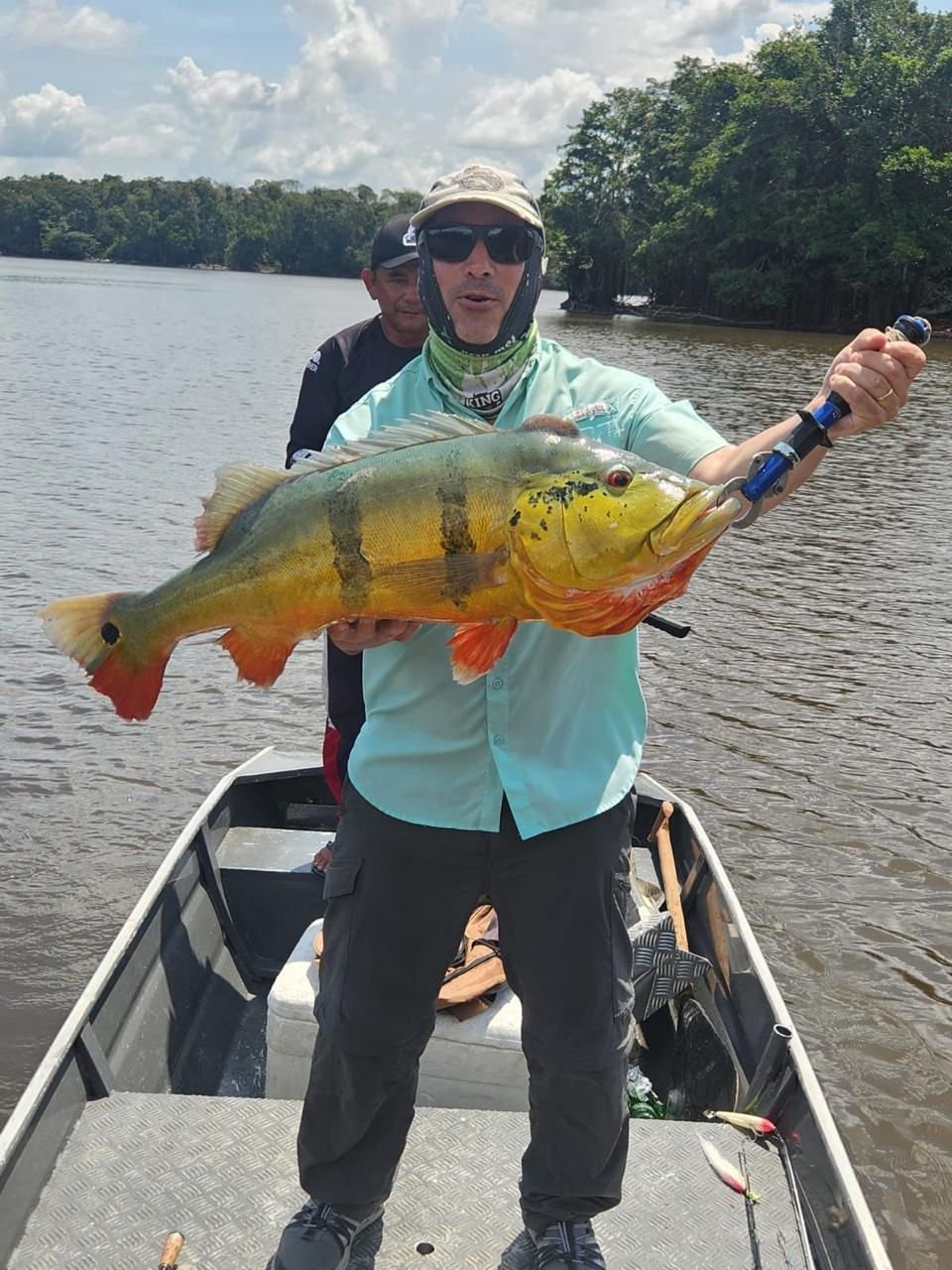 Man holding a large yellow and green fish with black vertical stripes on a boat in a river, with another person behind him and trees in the background.