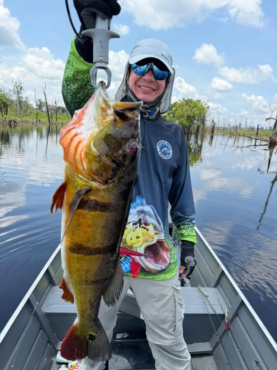 Person in a boat holding a large fish they caught, water, trees, and cloudy sky in the background.