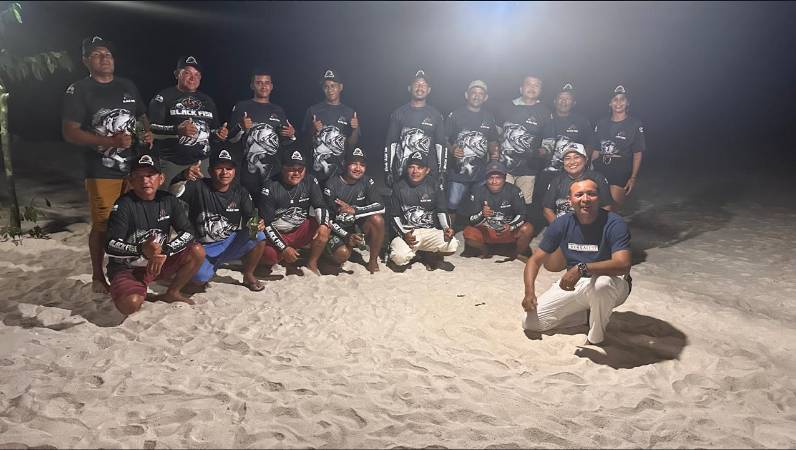 Group of people on a beach at night, wearing matching black shirts with a fish graphic and text, some are kneeling, others standing, posing for a group photo.
