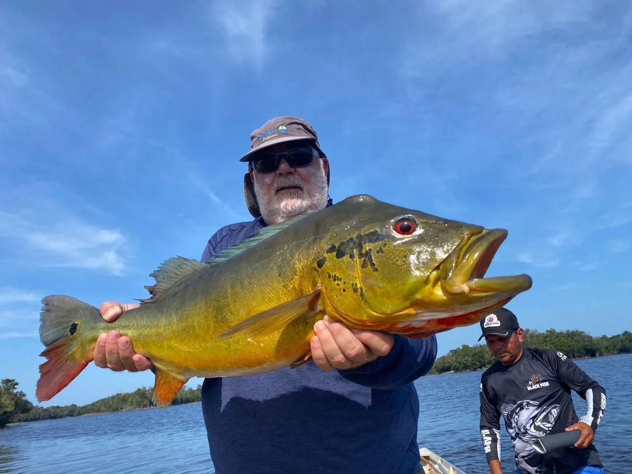 A man in sunglasses and a hat holding a large yellowish-green fish with an open mouth on a boat in a lake, with another man in a cap and fishing gear in the background.