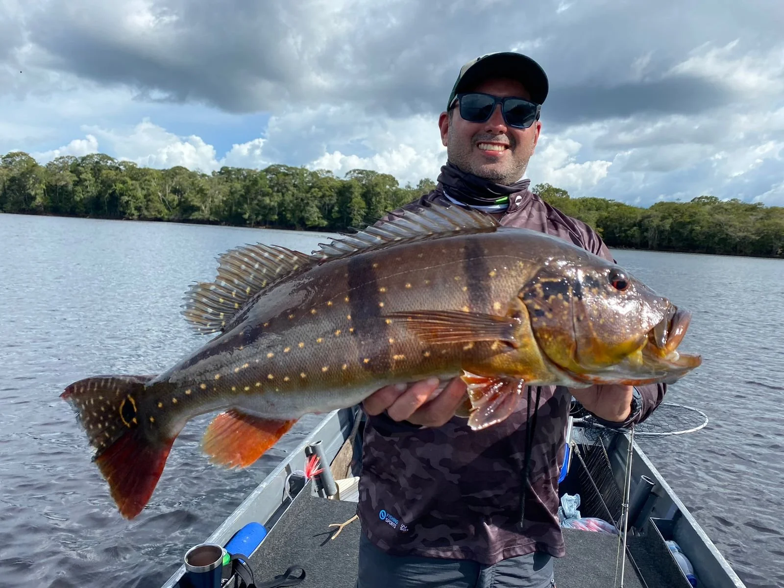 A smiling man wearing sunglasses and a cap holding a large fish over a body of water with trees in the background.