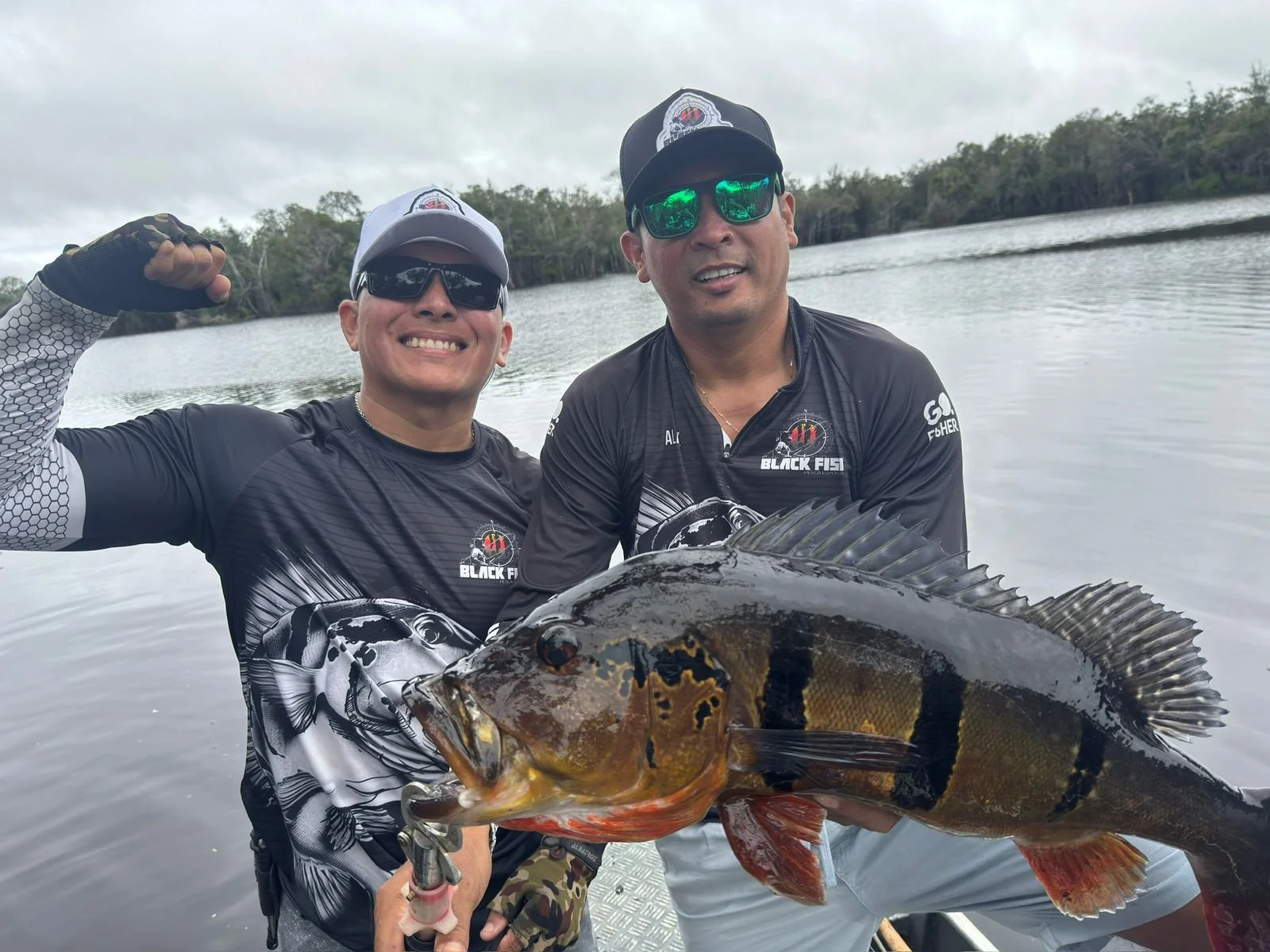 Two men on a boat holding a large fish they caught, with water and trees in the background.